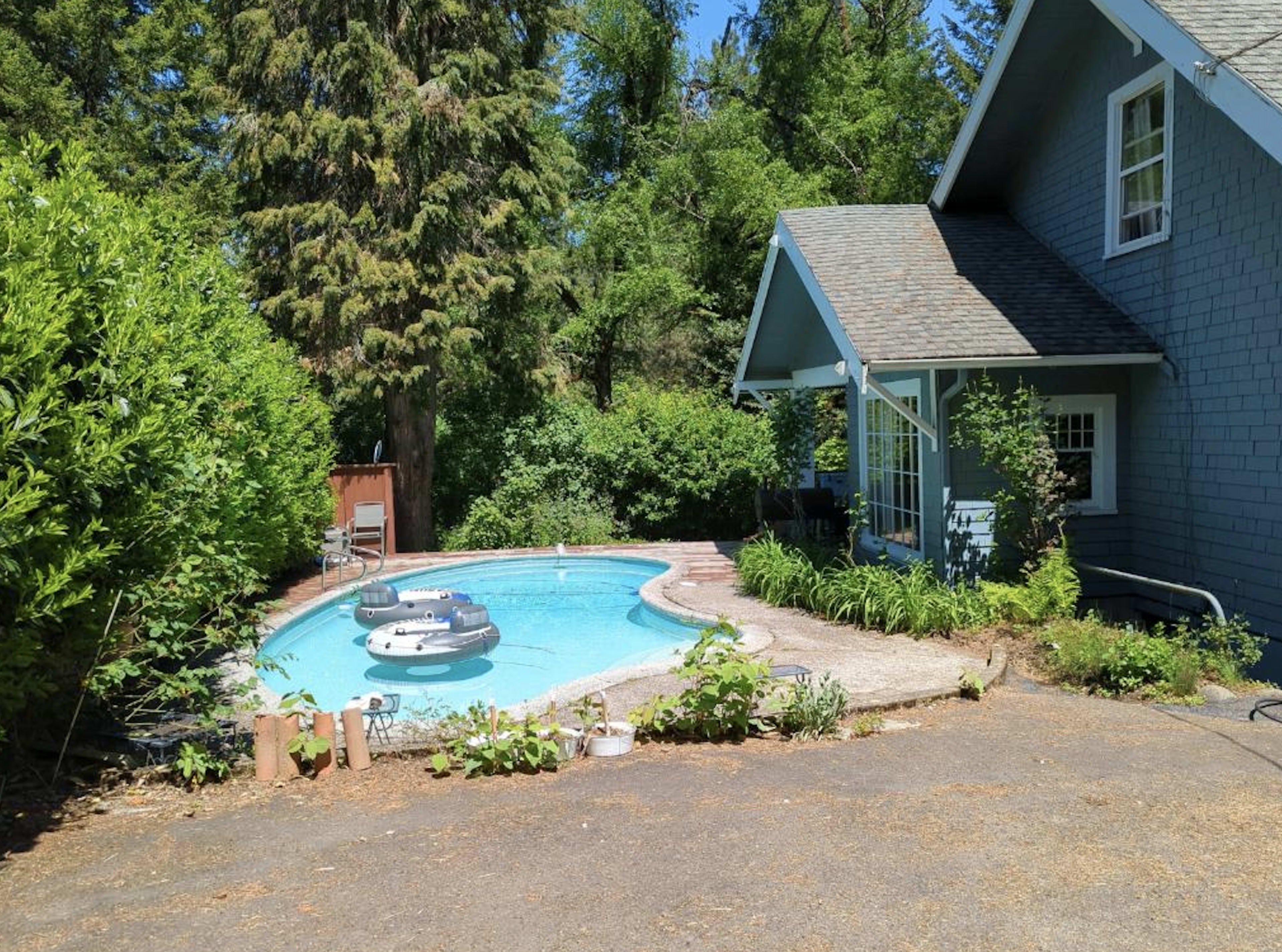 A blue house with a porch is situated next to a swimming pool surrounded by trees and greenery.