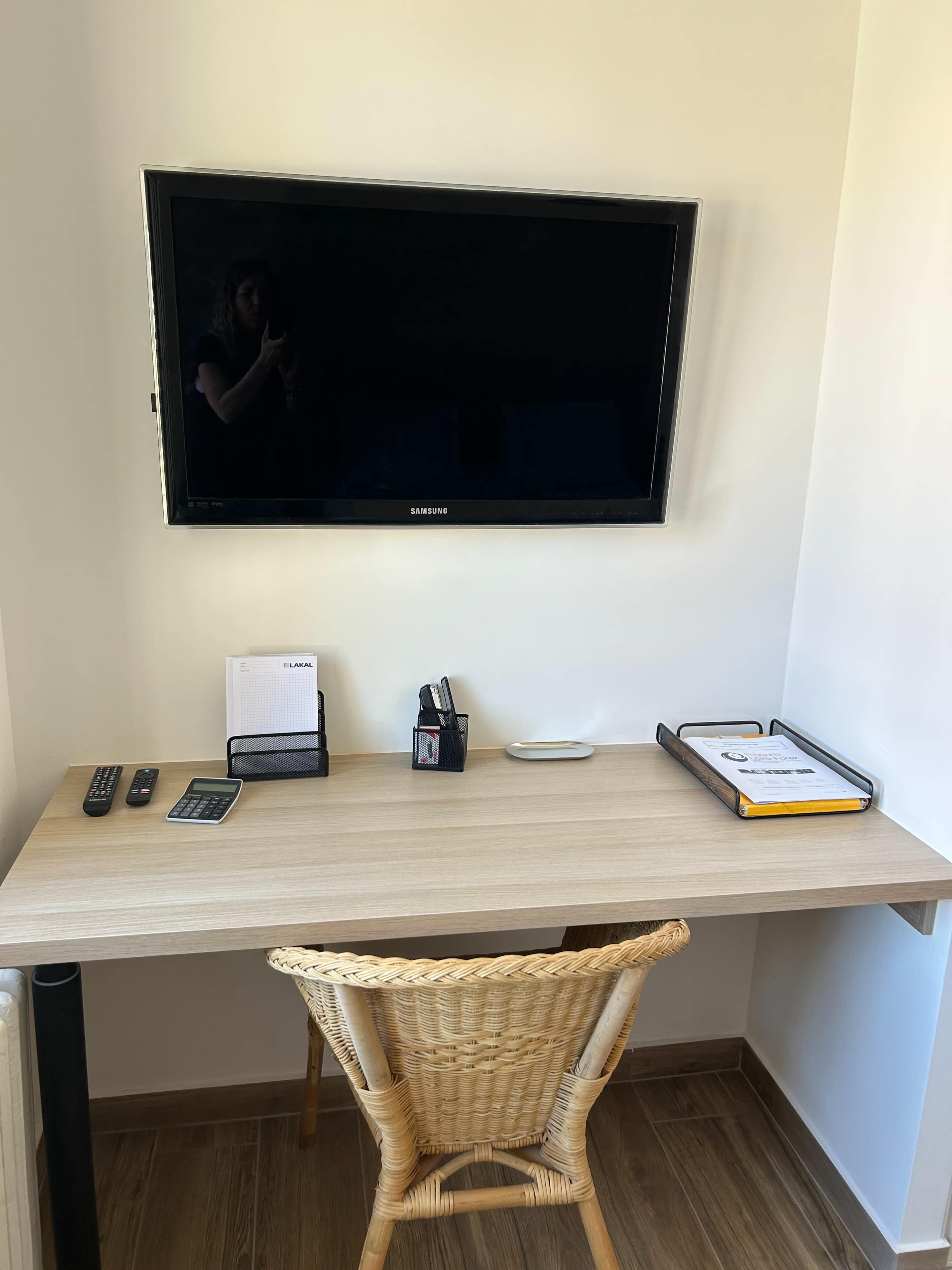 A wooden desk with a chair is positioned against a wall featuring a mounted television, along with remote controls and a folder organizer.