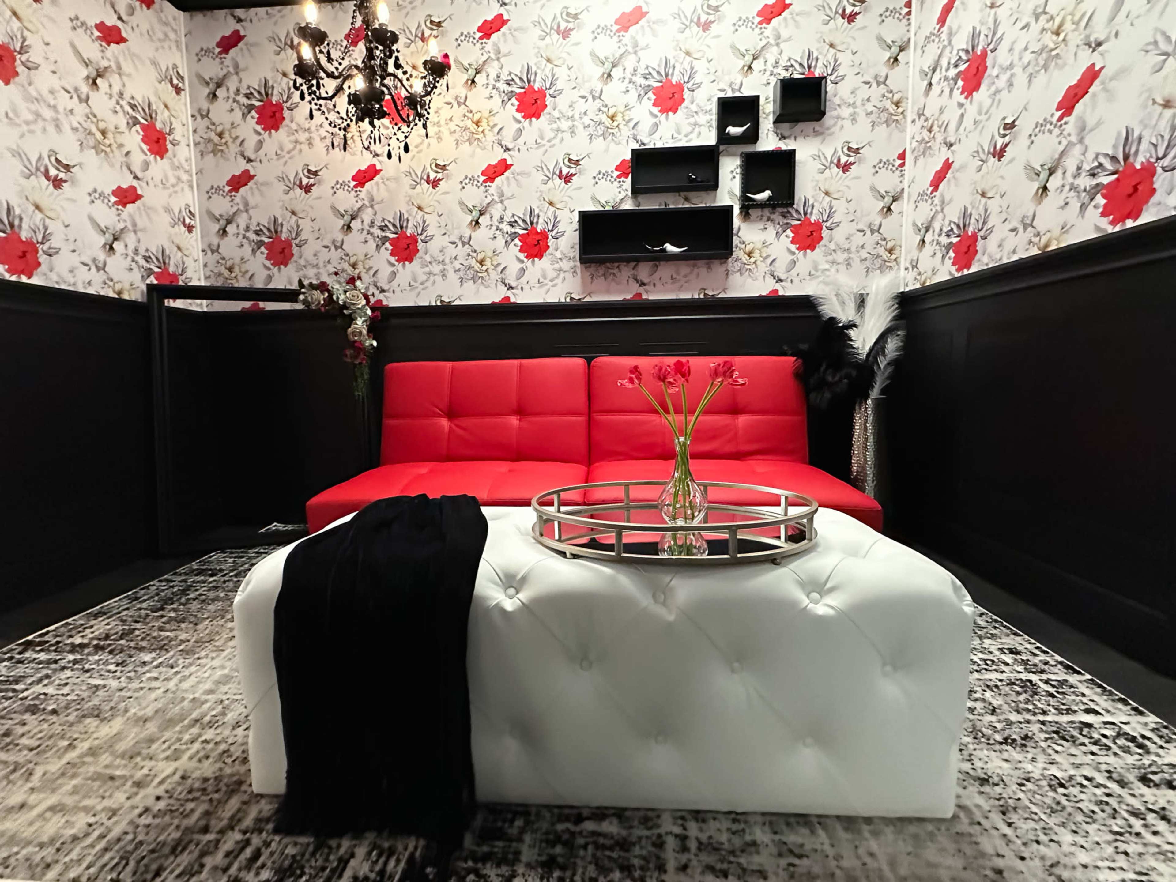 The image shows a room with black walls decorated with floral wallpaper, featuring a red sofa, a white ottoman, and a coffee table with a vase of flowers.