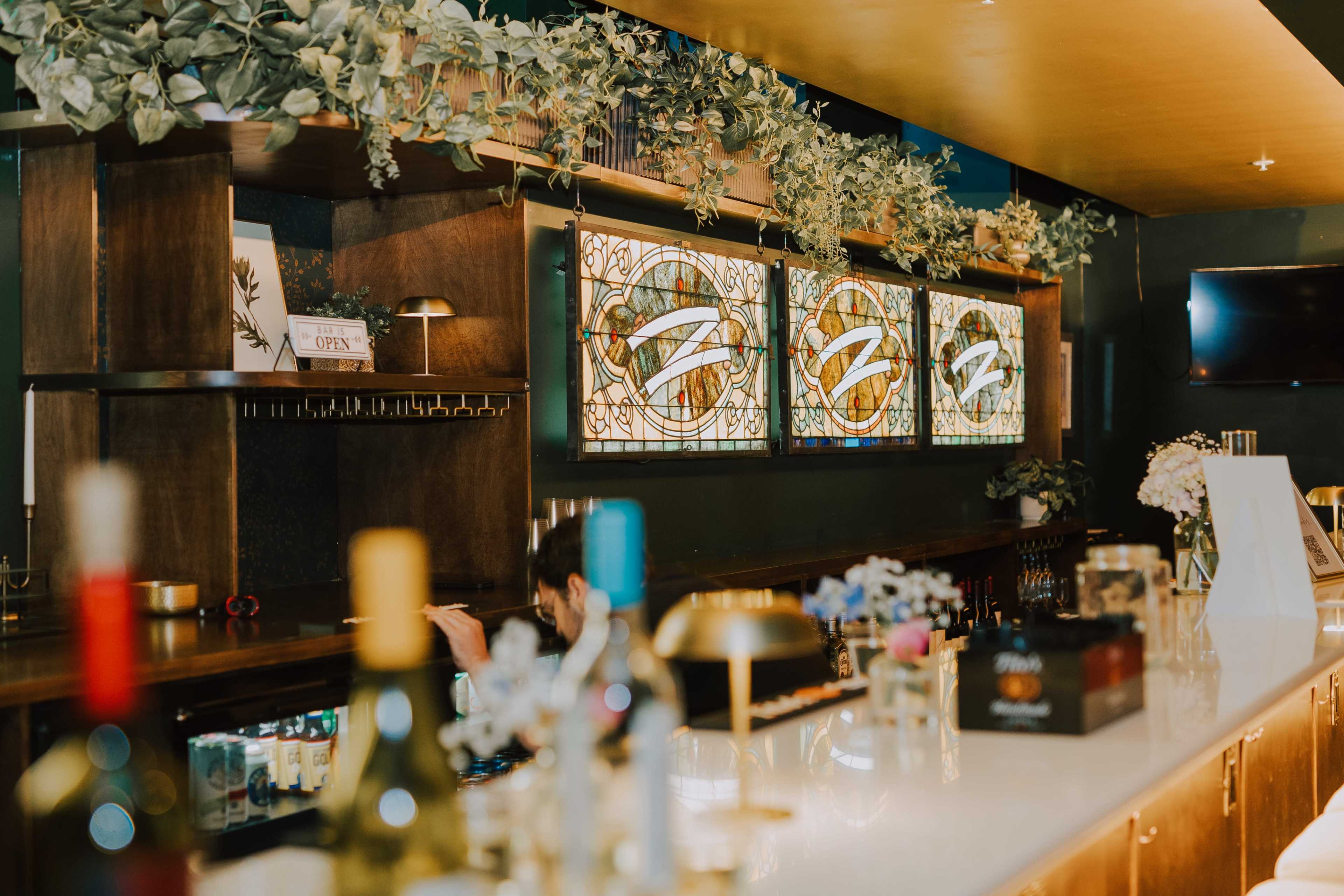 The image shows a bar area with a neatly arranged counter, decorative glass panels, and greenery hanging overhead.
