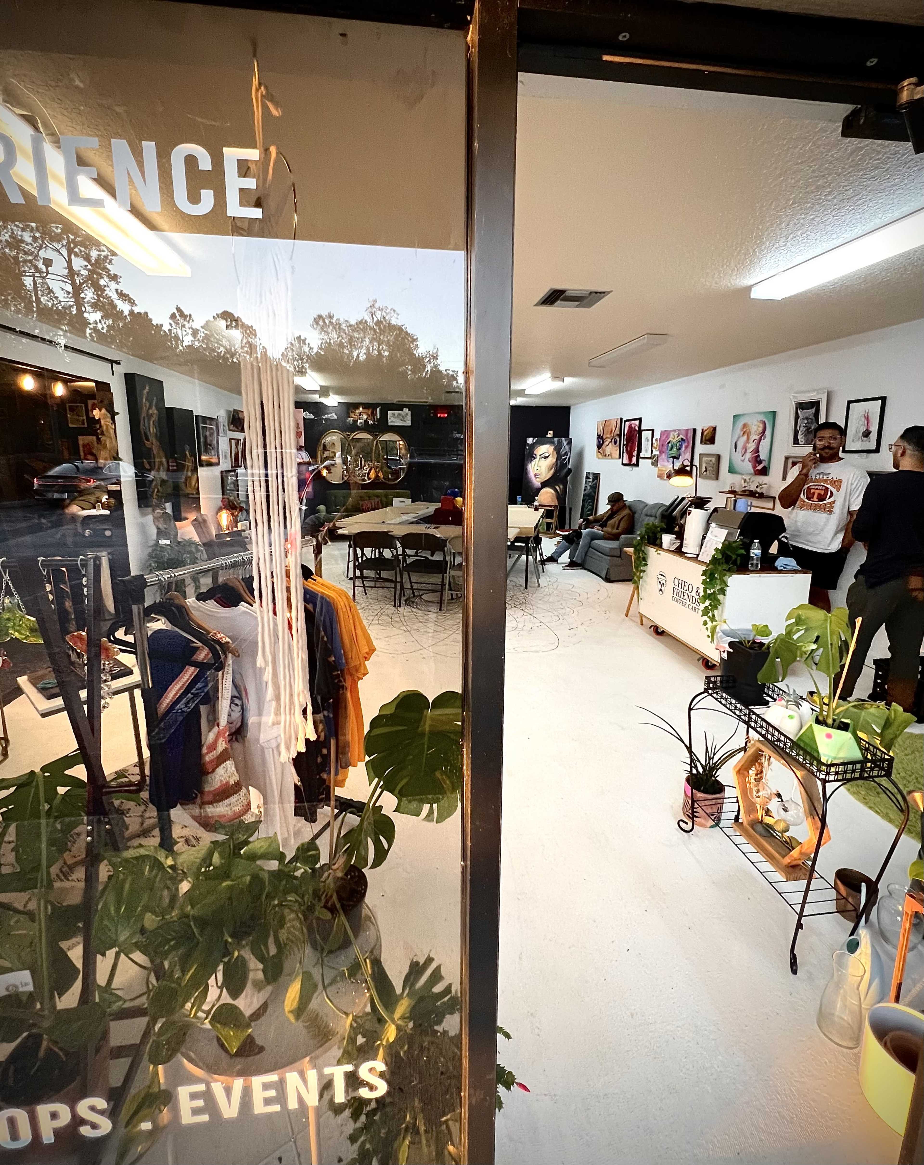 A view through a glass door into a creatively arranged indoor space featuring clothing displays, art on the walls, and tables set up for an event.