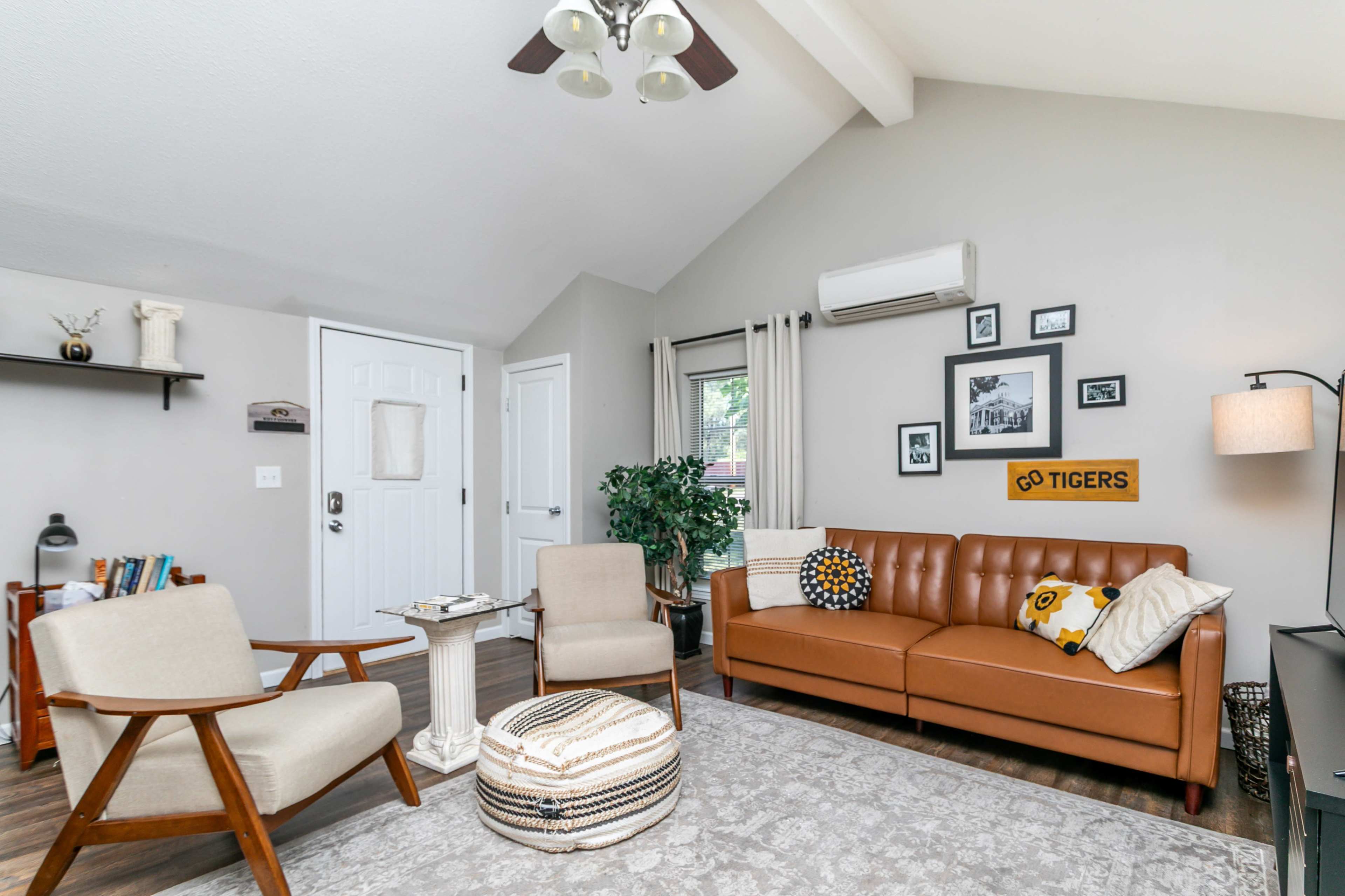 The living room features a brown leather sofa, two armchairs, a coffee table, a potted plant, and framed artwork on the walls, along with a door leading outside.