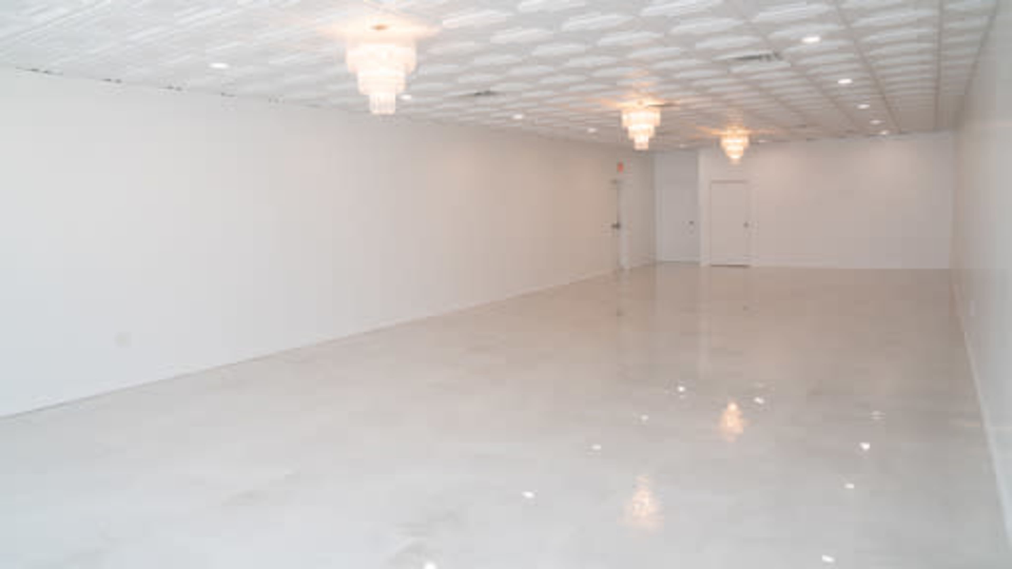 The image shows a spacious, empty room with white walls and a glossy floor, featuring ceiling lights and two chandeliers.
