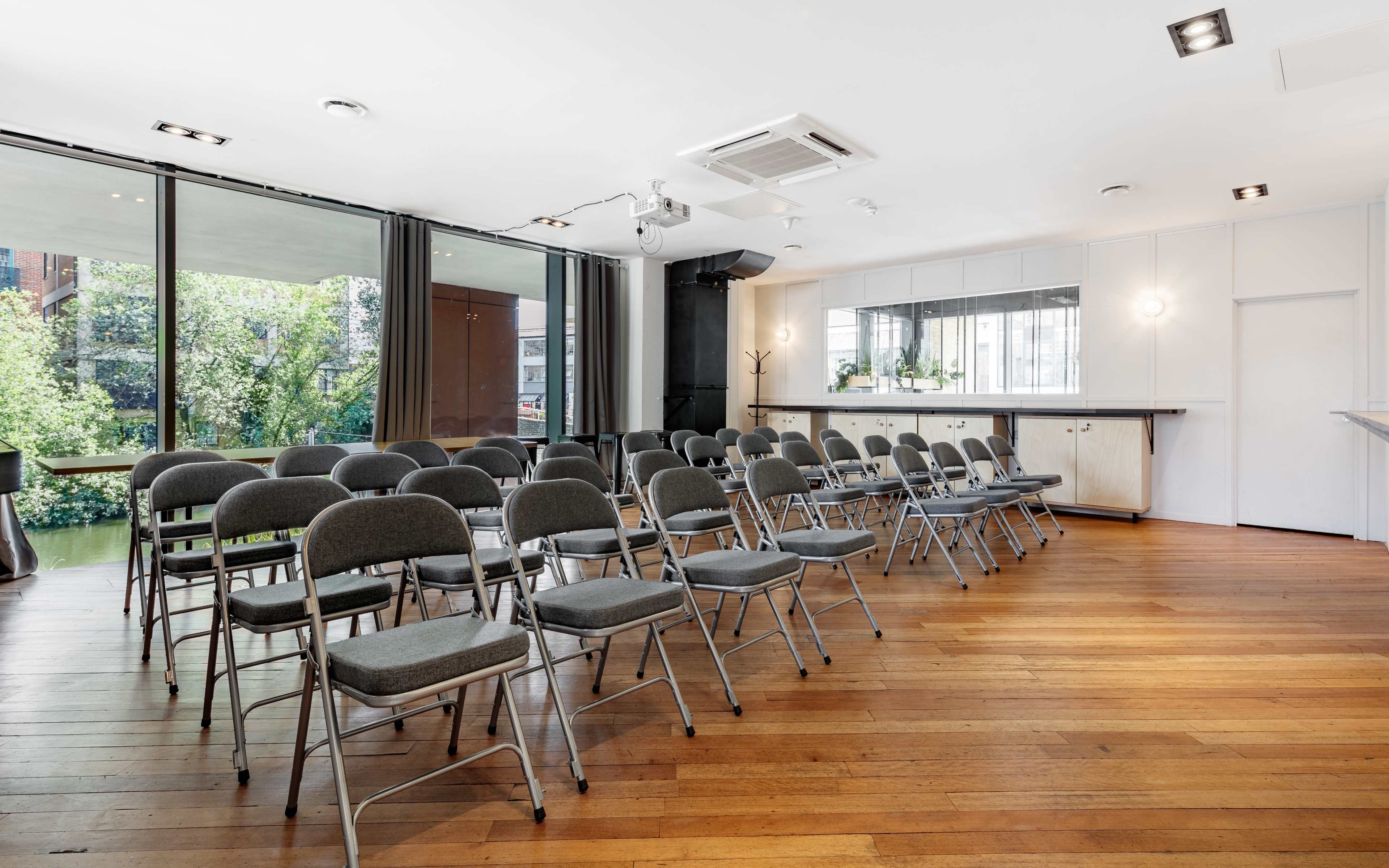 A modern meeting room with rows of metal folding chairs arranged neatly on a wooden floor, overlooking a green outdoor area through large windows.