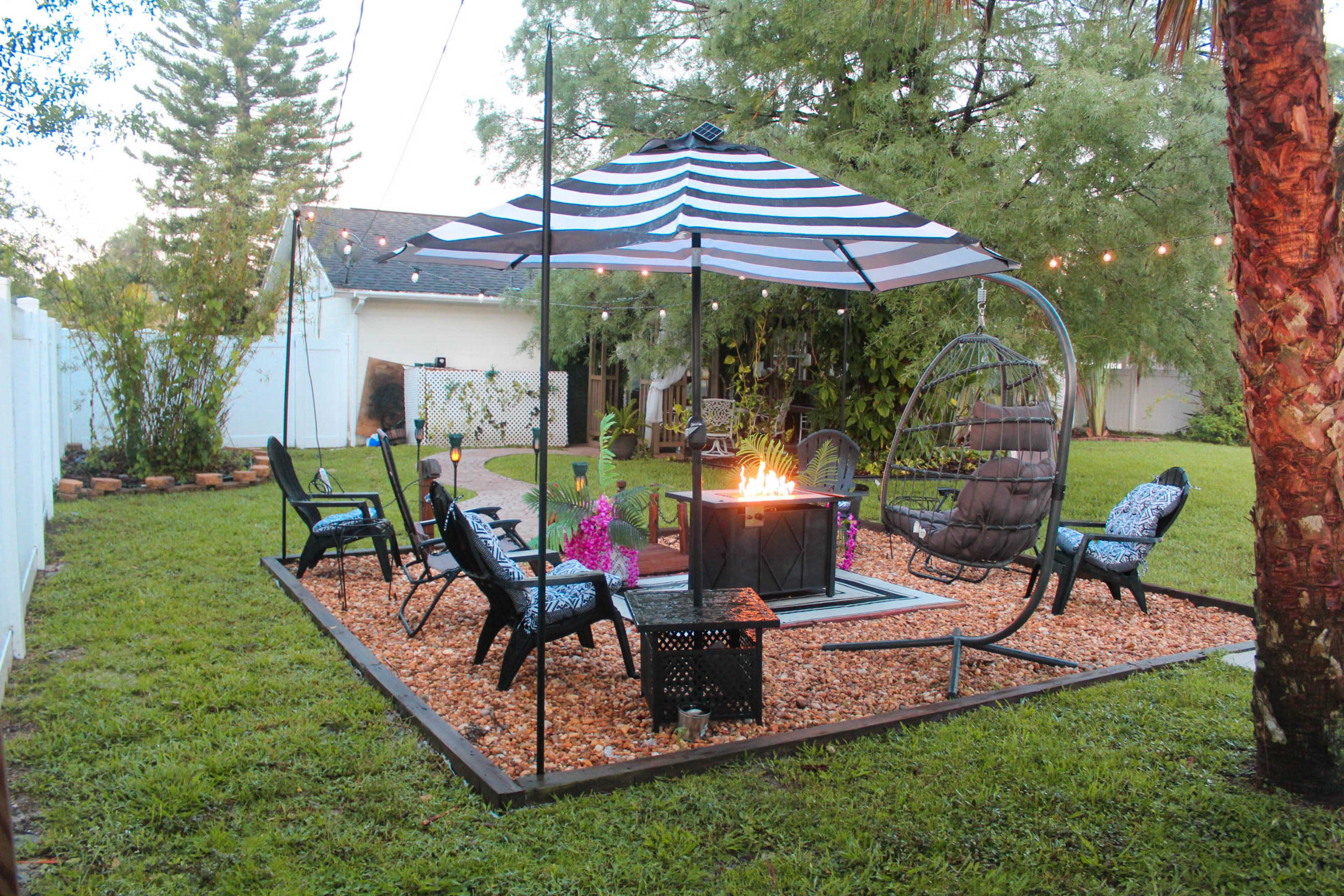 A backyard seating area features a fire pit surrounded by chairs, a swinging chair, and a striped canopy, all set on gravel and grass.