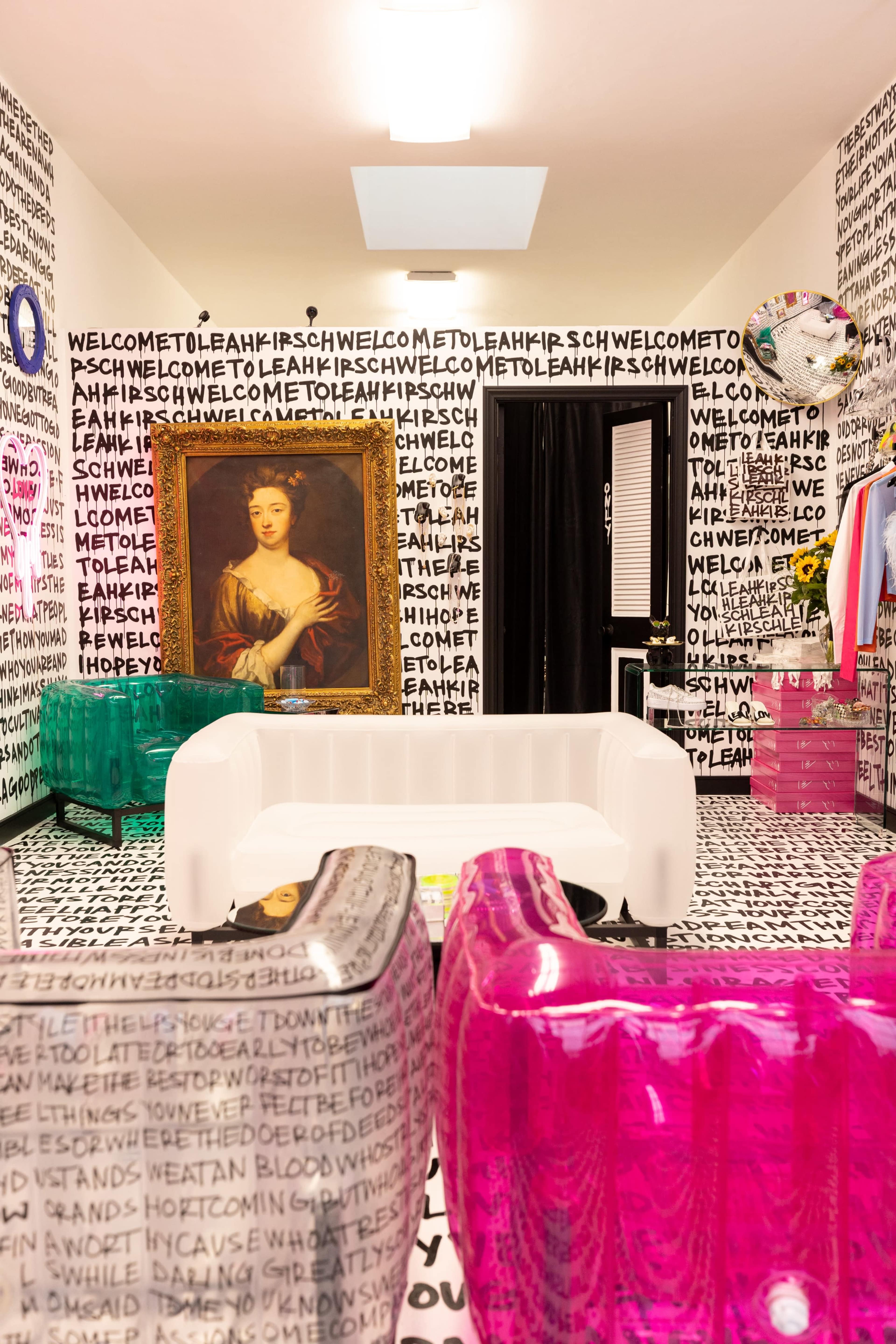 A vividly designed room features transparent colored furniture, a large golden-framed portrait, and walls covered in repetitive text patterns.