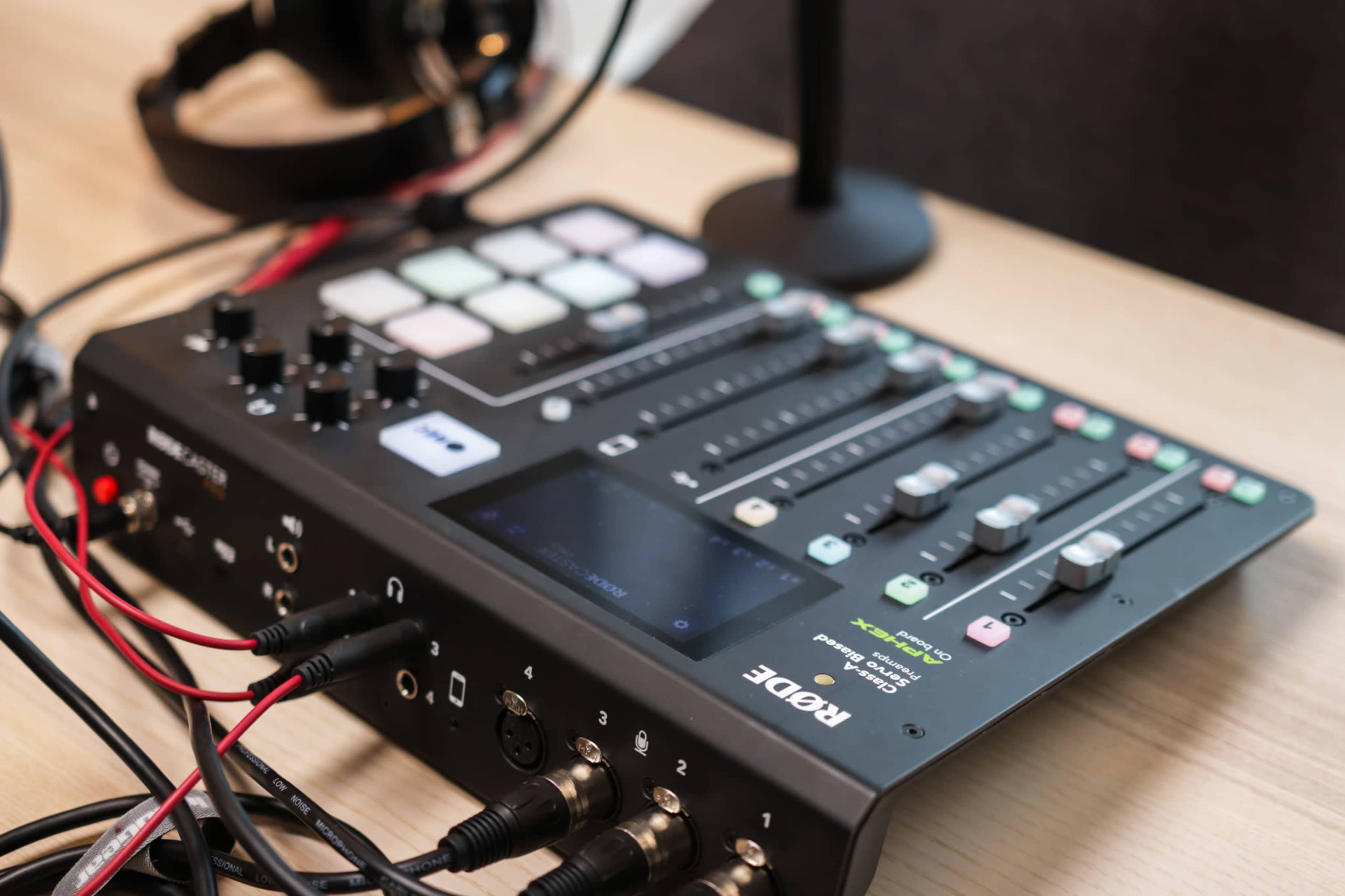 A Rode audio mixer featuring multiple sliders, buttons, and audio input/output connections is displayed on a wooden surface.