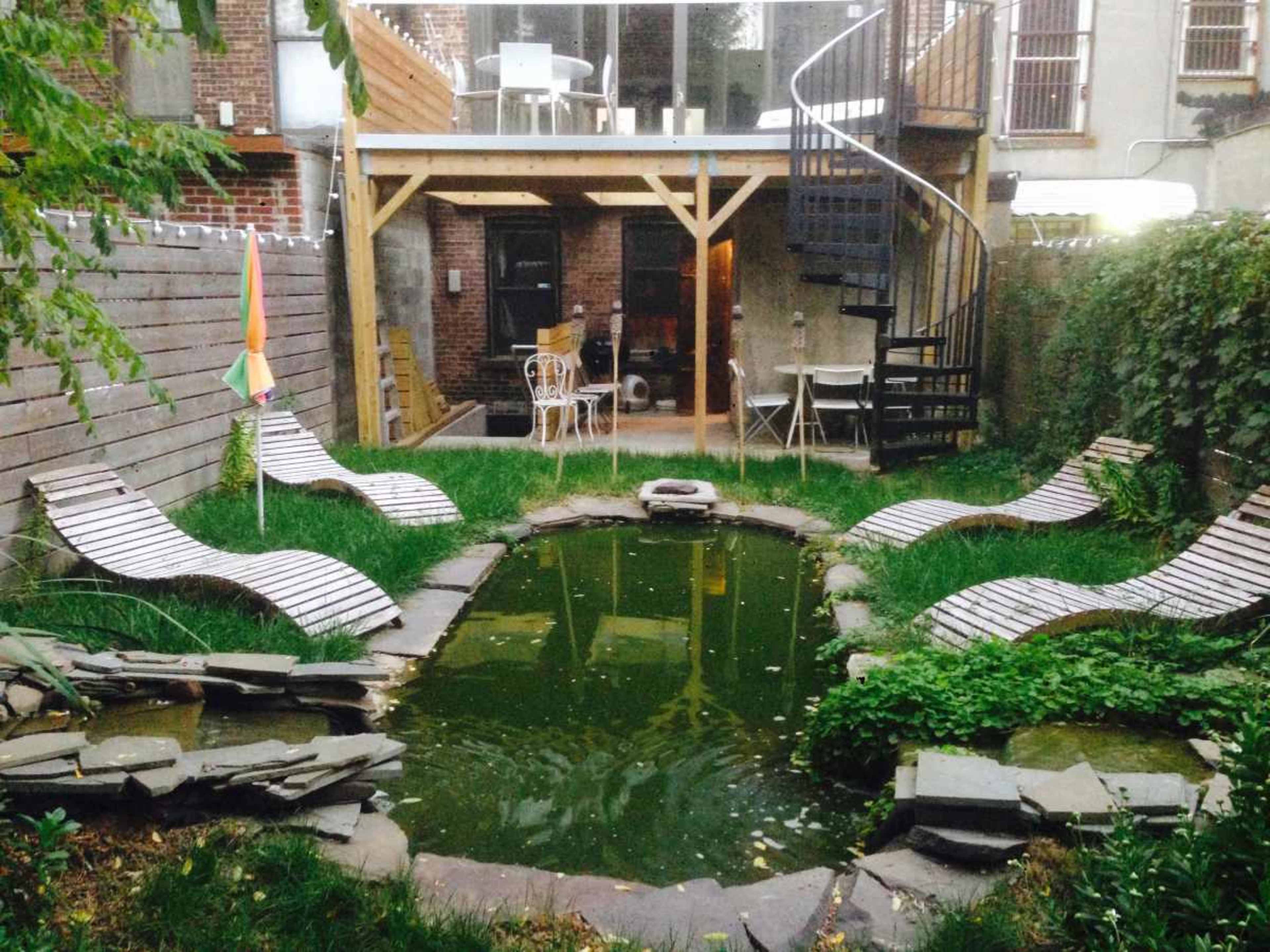 The image shows a small outdoor garden area with a round pond at the center, surrounded by wooden lounge chairs and a two-story building in the background.