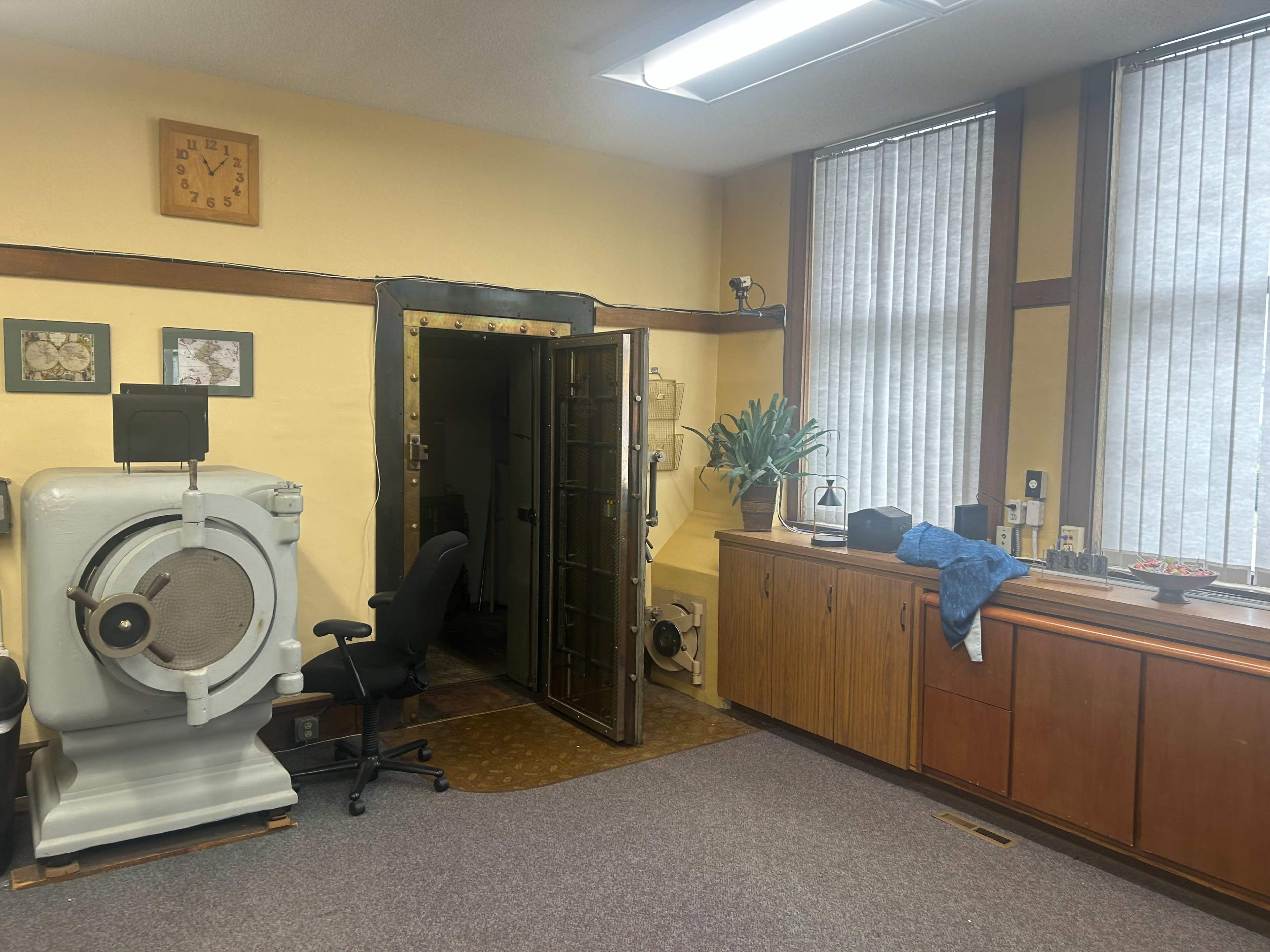 The image displays a room with a large, vintage vault door and a professional chair, accompanied by a wooden cabinet and a wall clock.