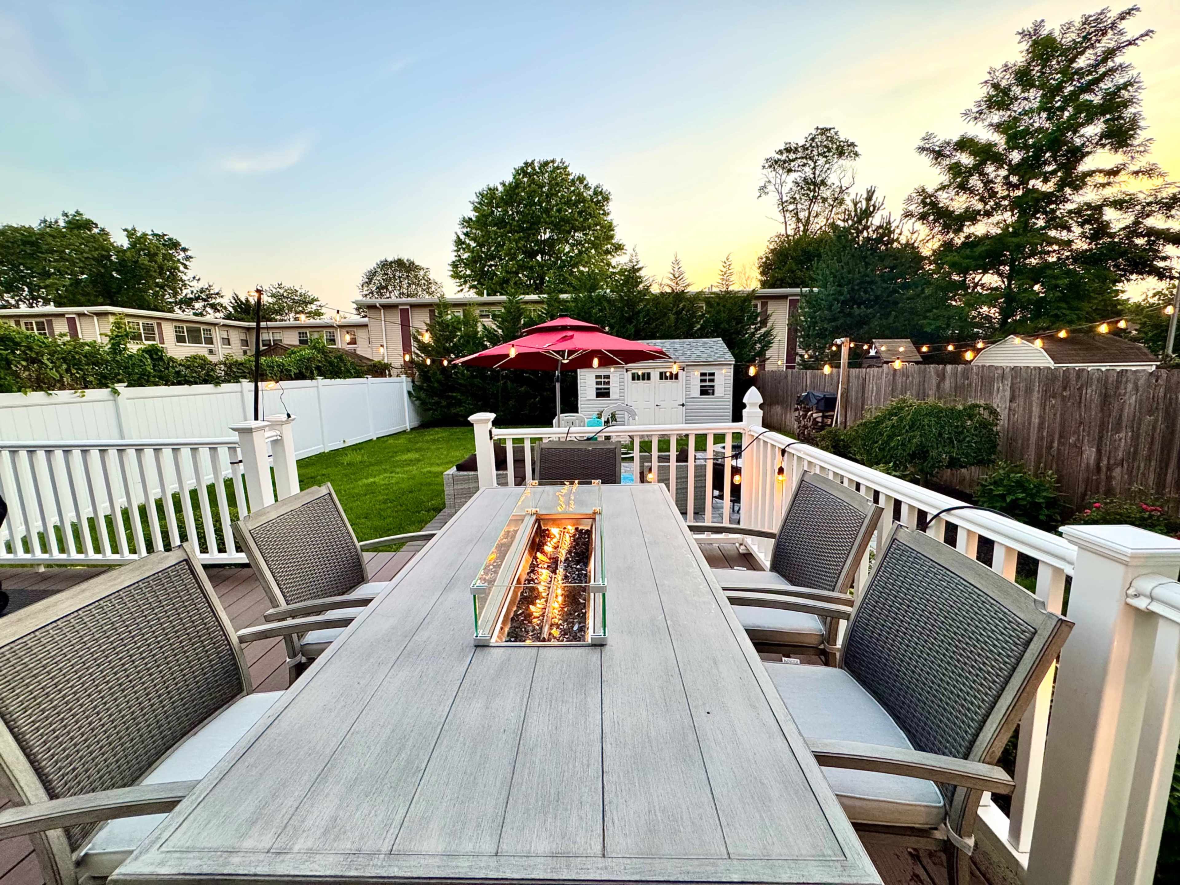 A wooden dining table with a central fire feature is set on a deck surrounded by chairs, with a green lawn and a gazebo in the background under a sunset sky.