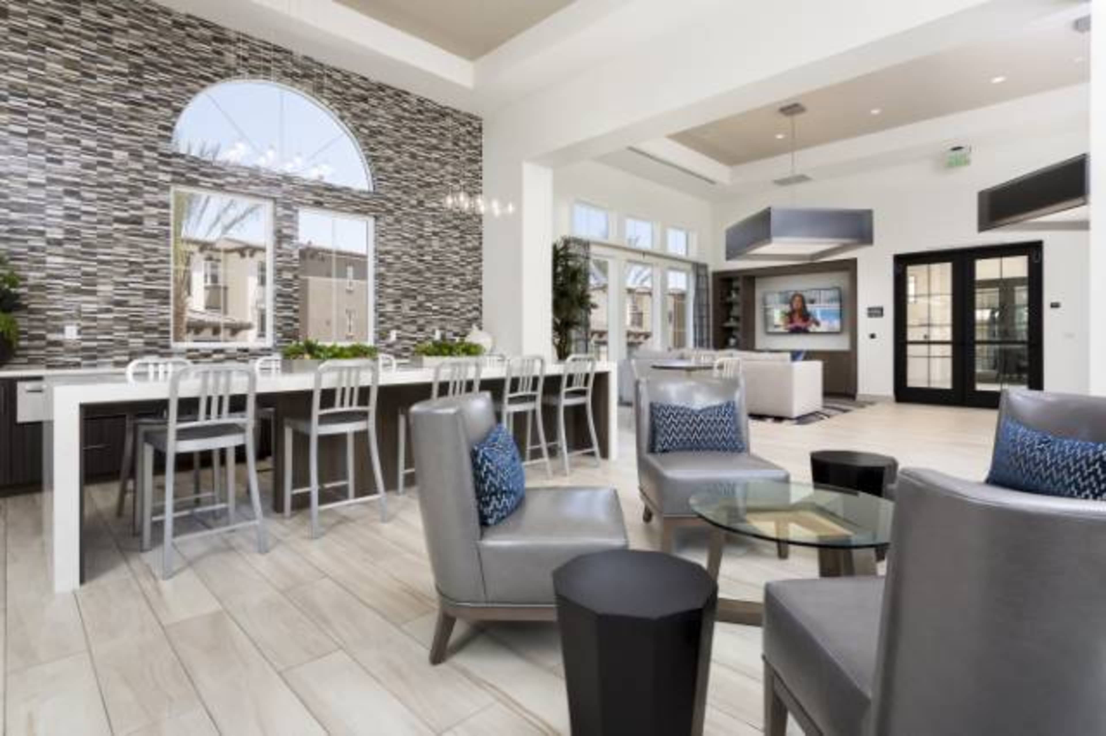 A modern indoor space featuring a kitchen bar with stools, a seating area with gray chairs, and large windows allowing natural light to enter.