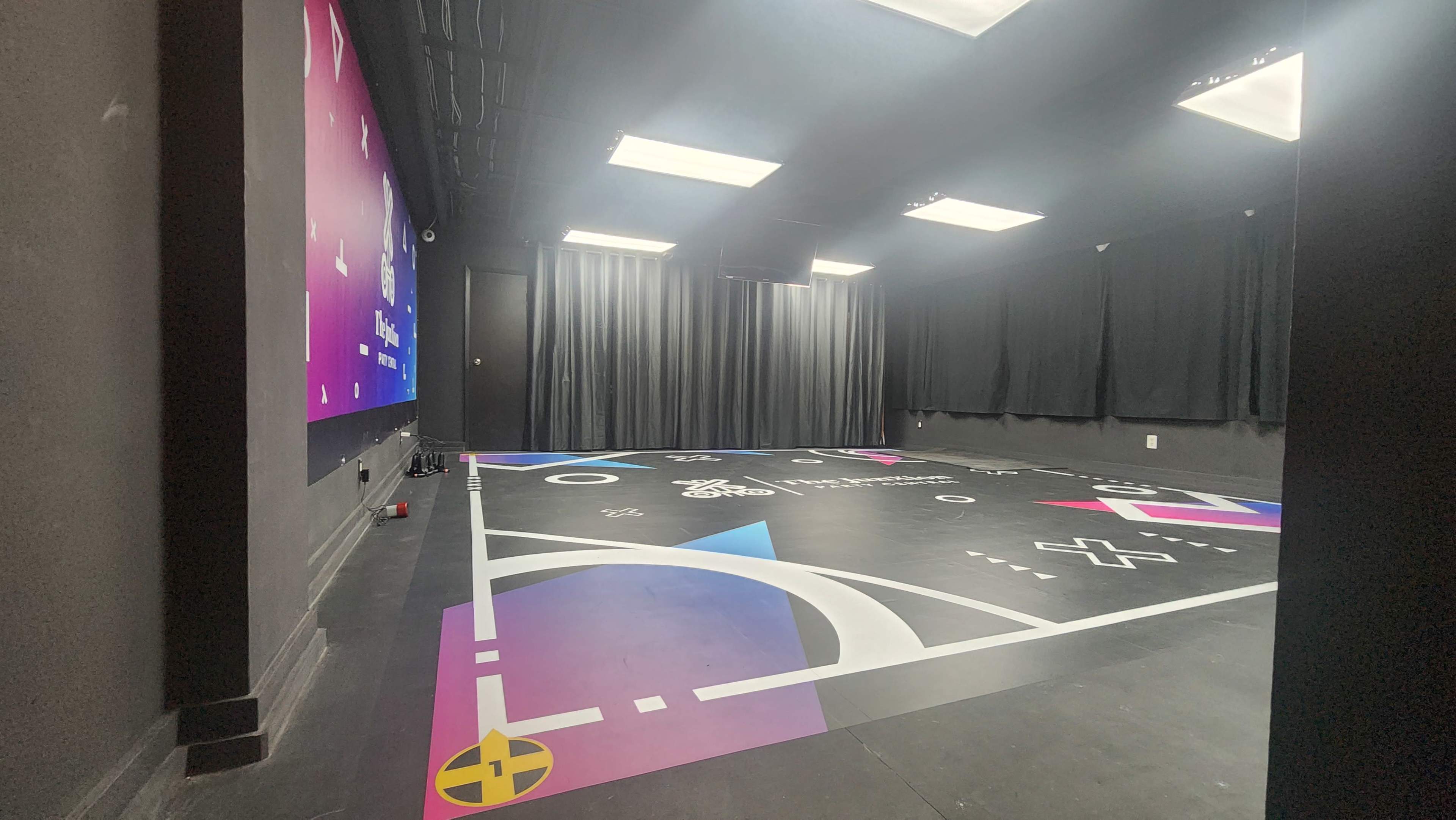 The image shows a brightly colored gym or studio space with a patterned floor, black curtains, and a colorful wall mural.