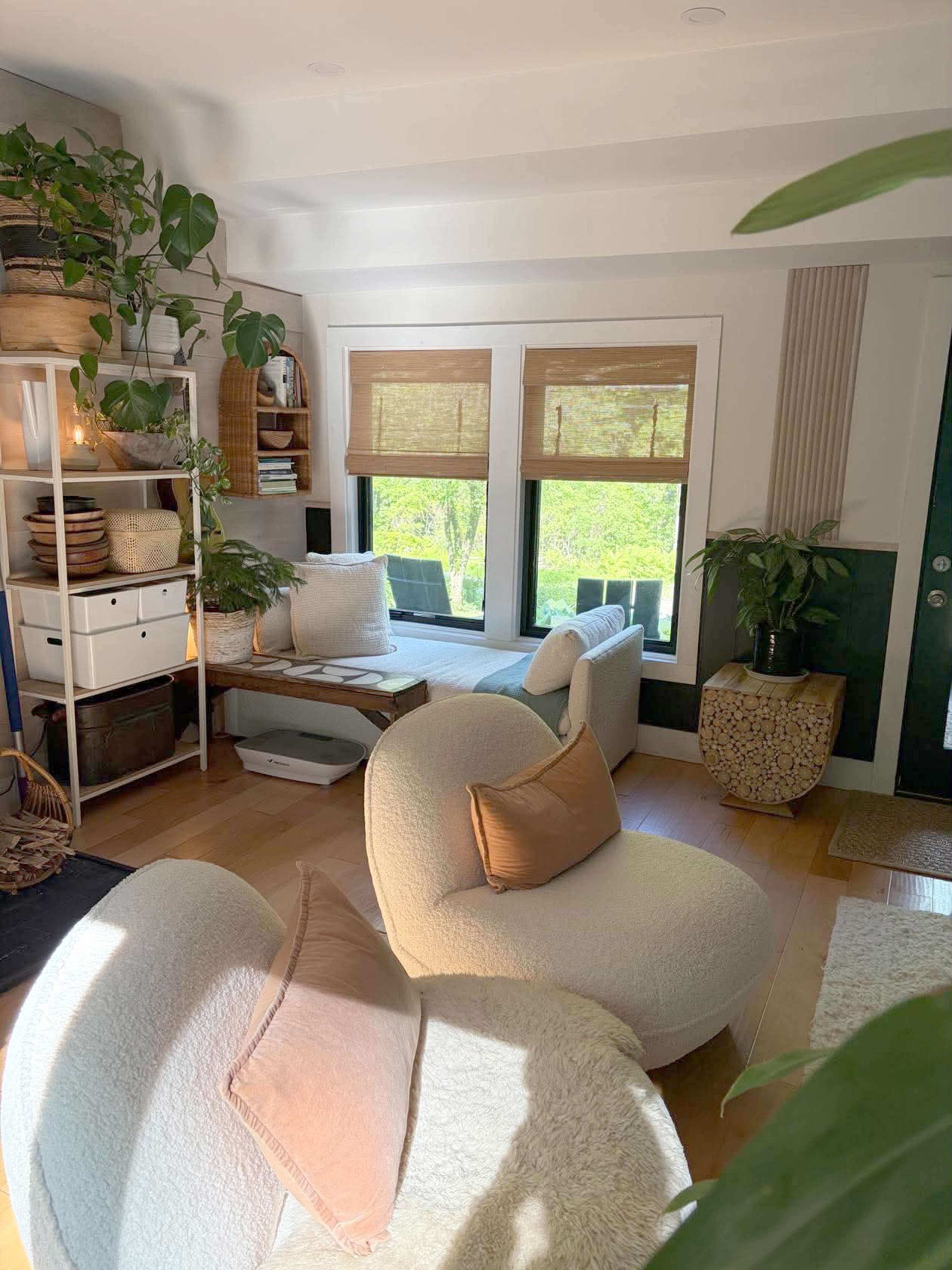 The image shows a cozy living room with a curved white sofa, large windows with bamboo shades, and various indoor plants.