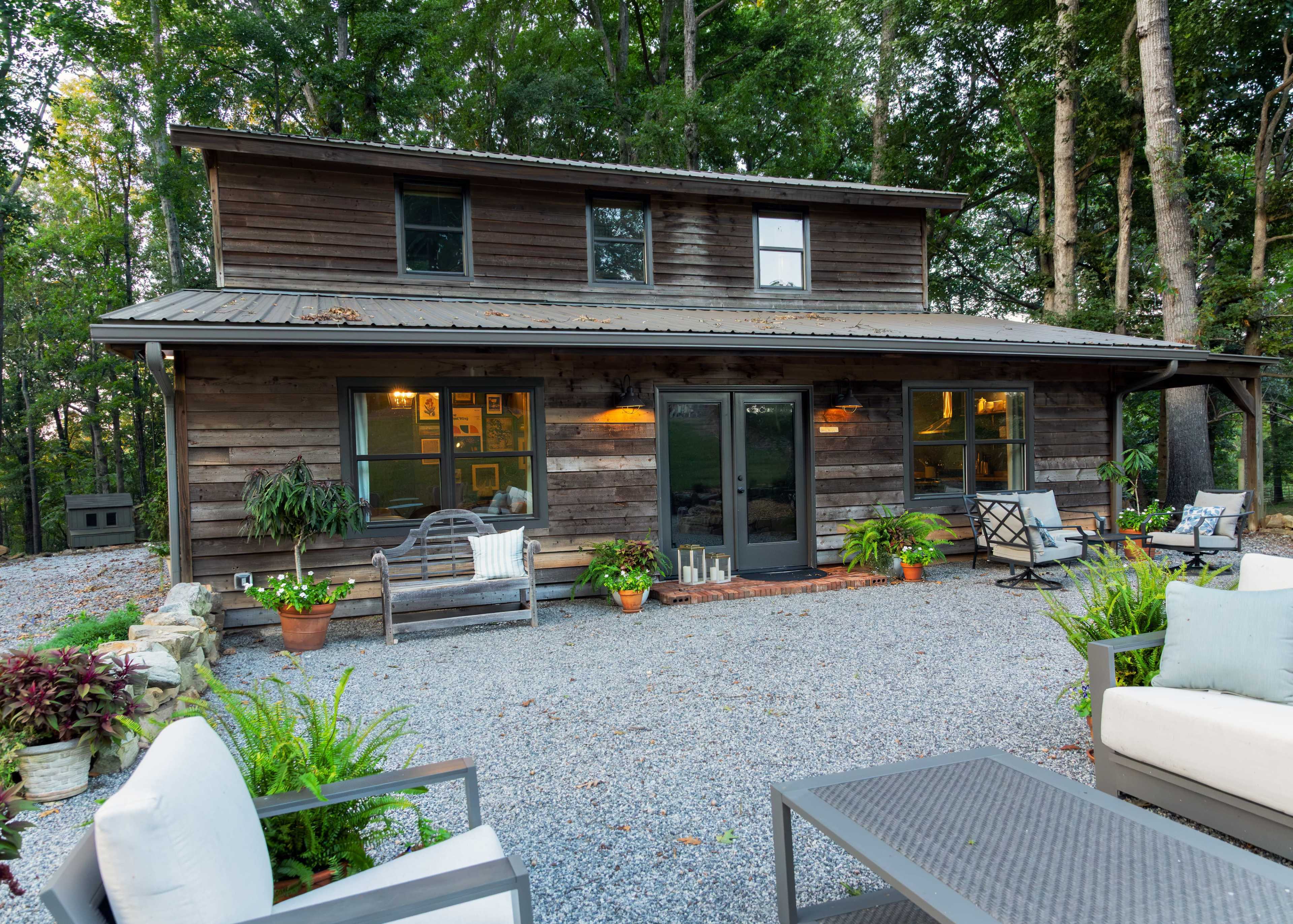A two-story wooden house with a metal roof is surrounded by dense trees and features a gravel patio with potted plants and outdoor seating.