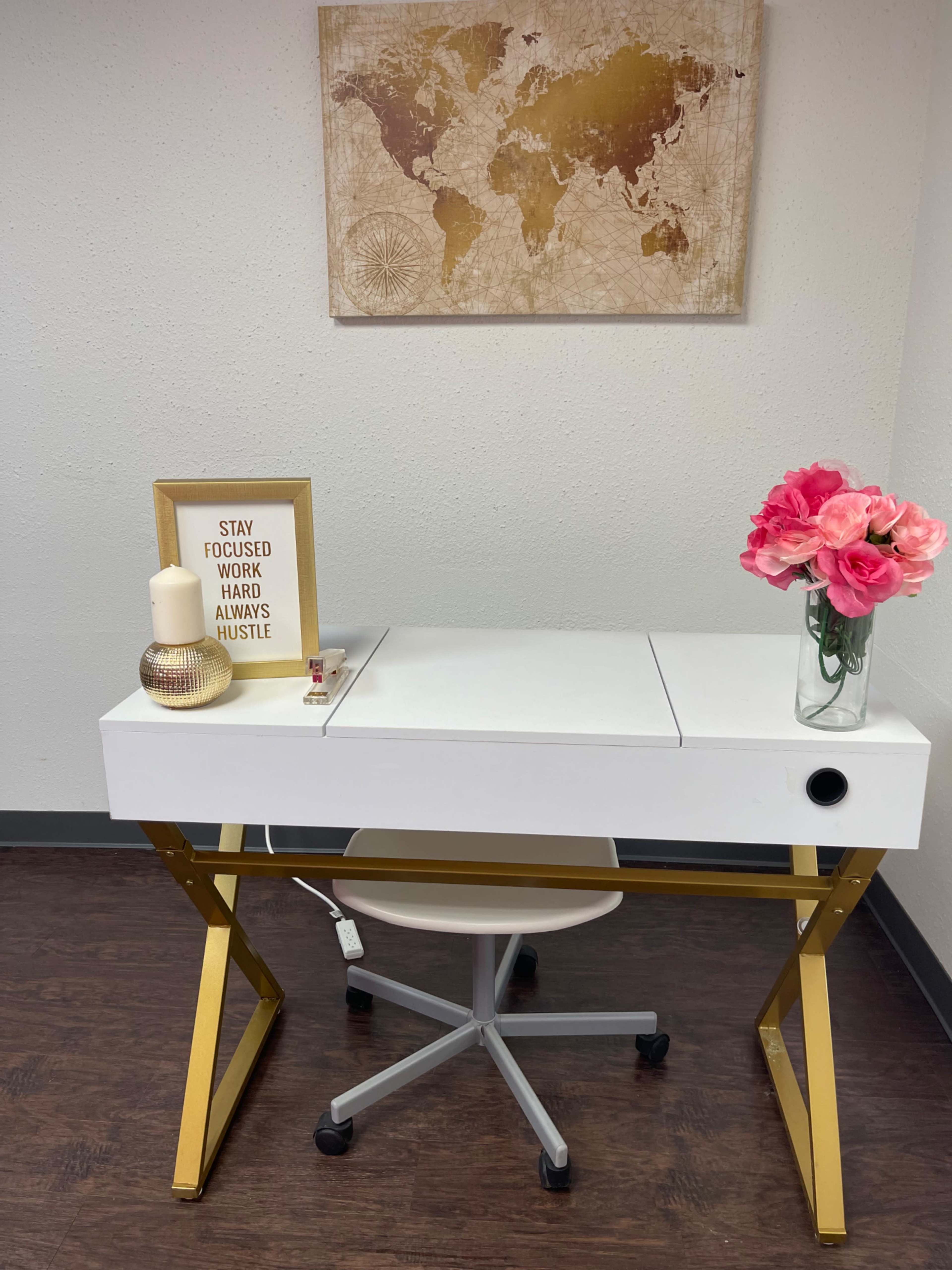 A white desk with gold legs is positioned against a wall, featuring a map art piece and decor items including a candle, a framed quote, and a vase of pink flowers.