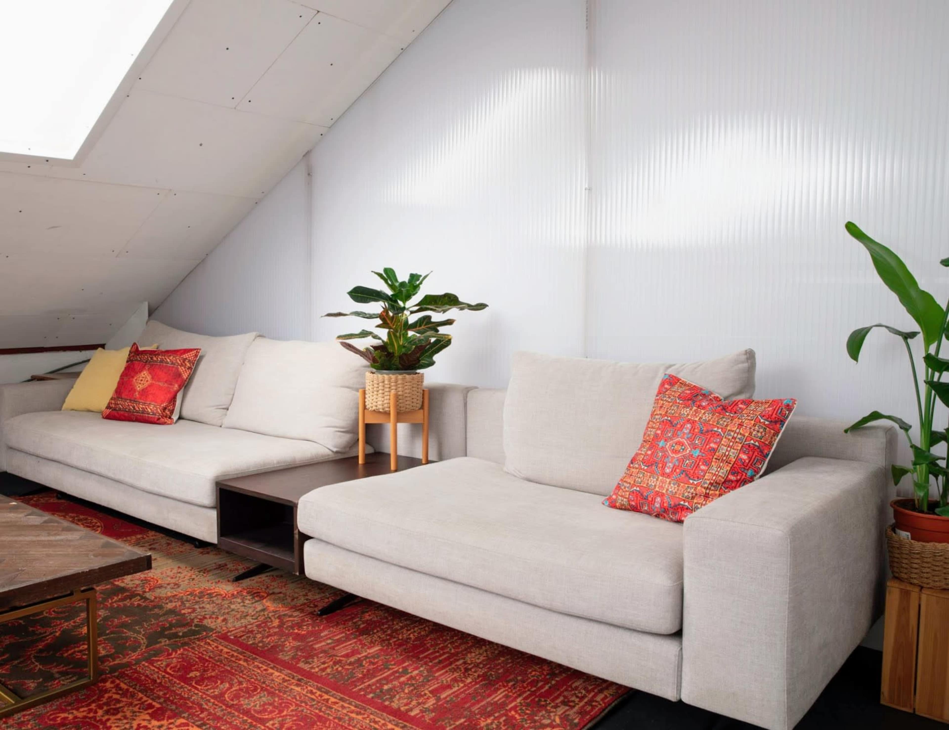 A cozy living area features two light-colored sofas with decorative pillows, a small wooden table, and indoor plants, set against a backdrop of angled walls.