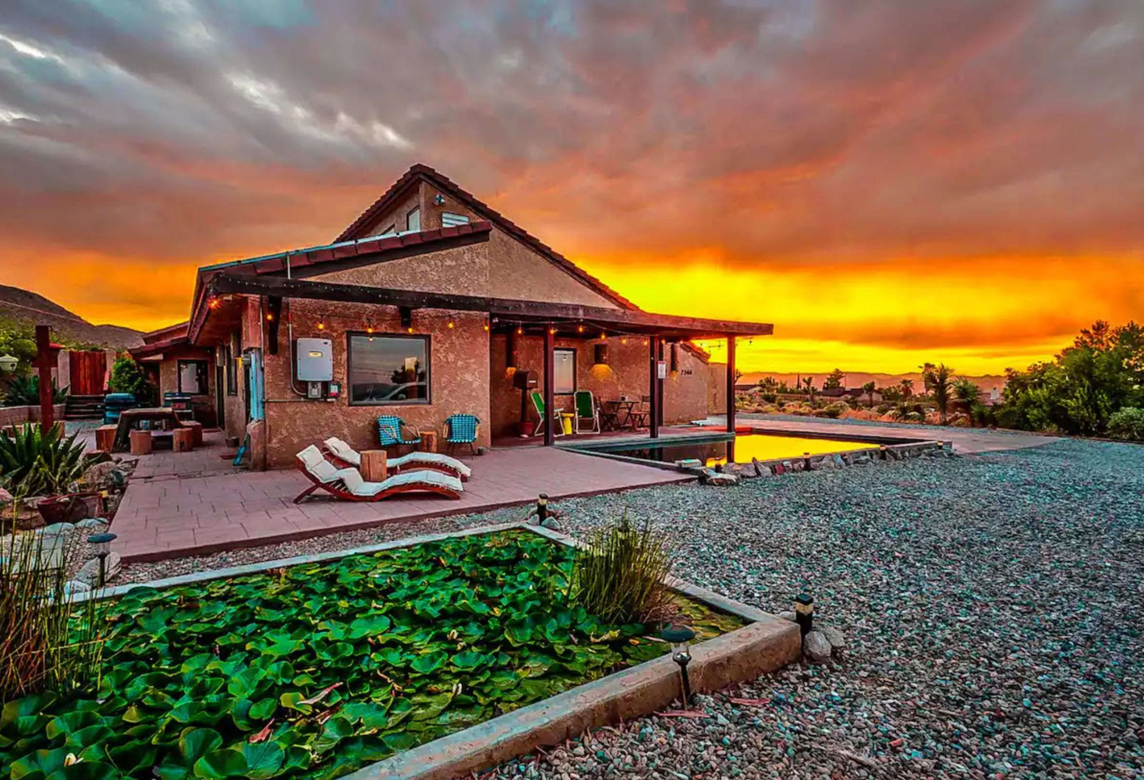 A house with a sloped roof is set against a vibrant sunset, with a pool and a landscaped area featuring lily pads in the foreground.