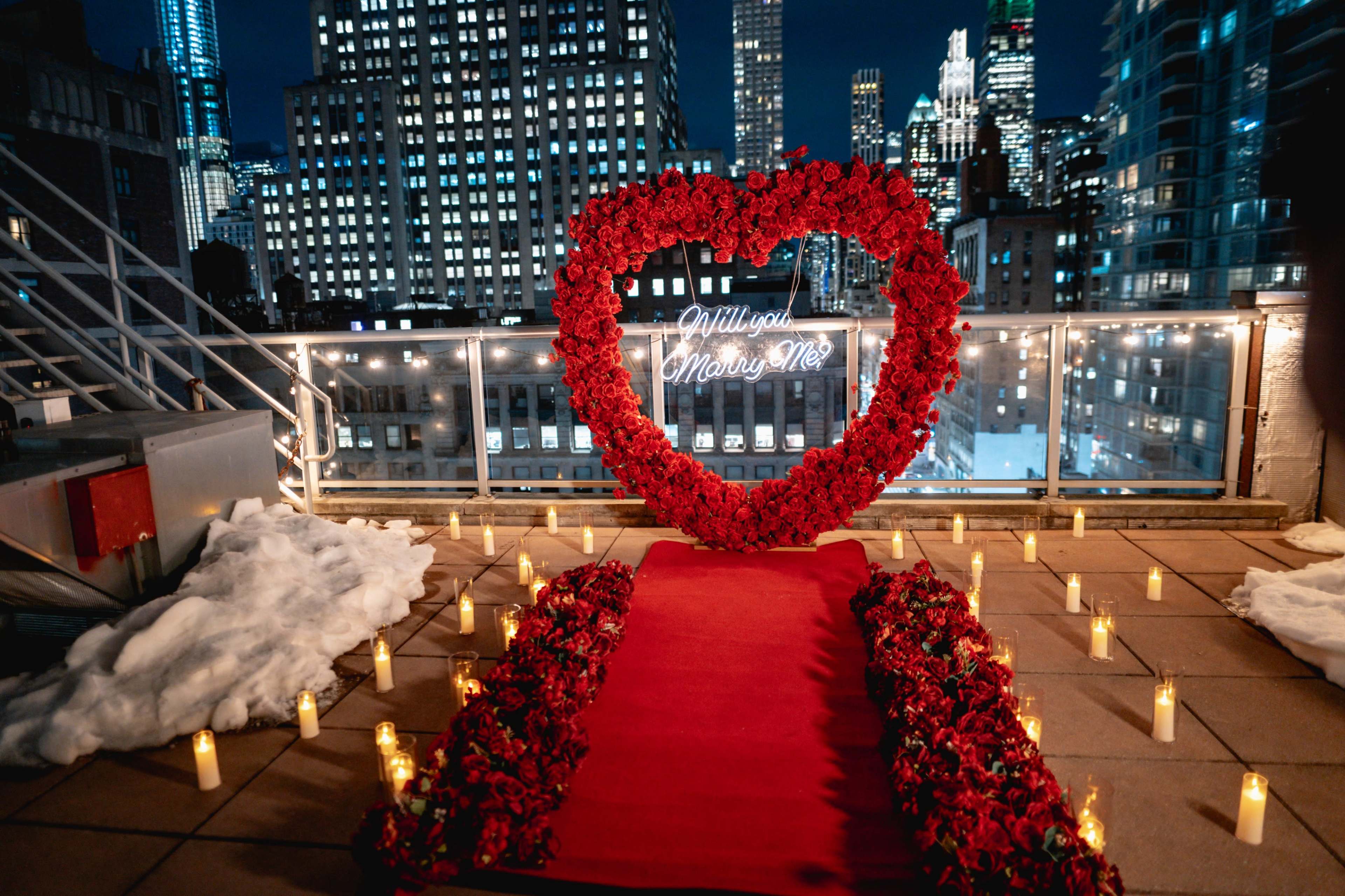 A heart-shaped floral arrangement with a neon sign reading "Will you marry me?" is set up on a rooftop terrace adorned with candles, overlooking a city skyline at night.