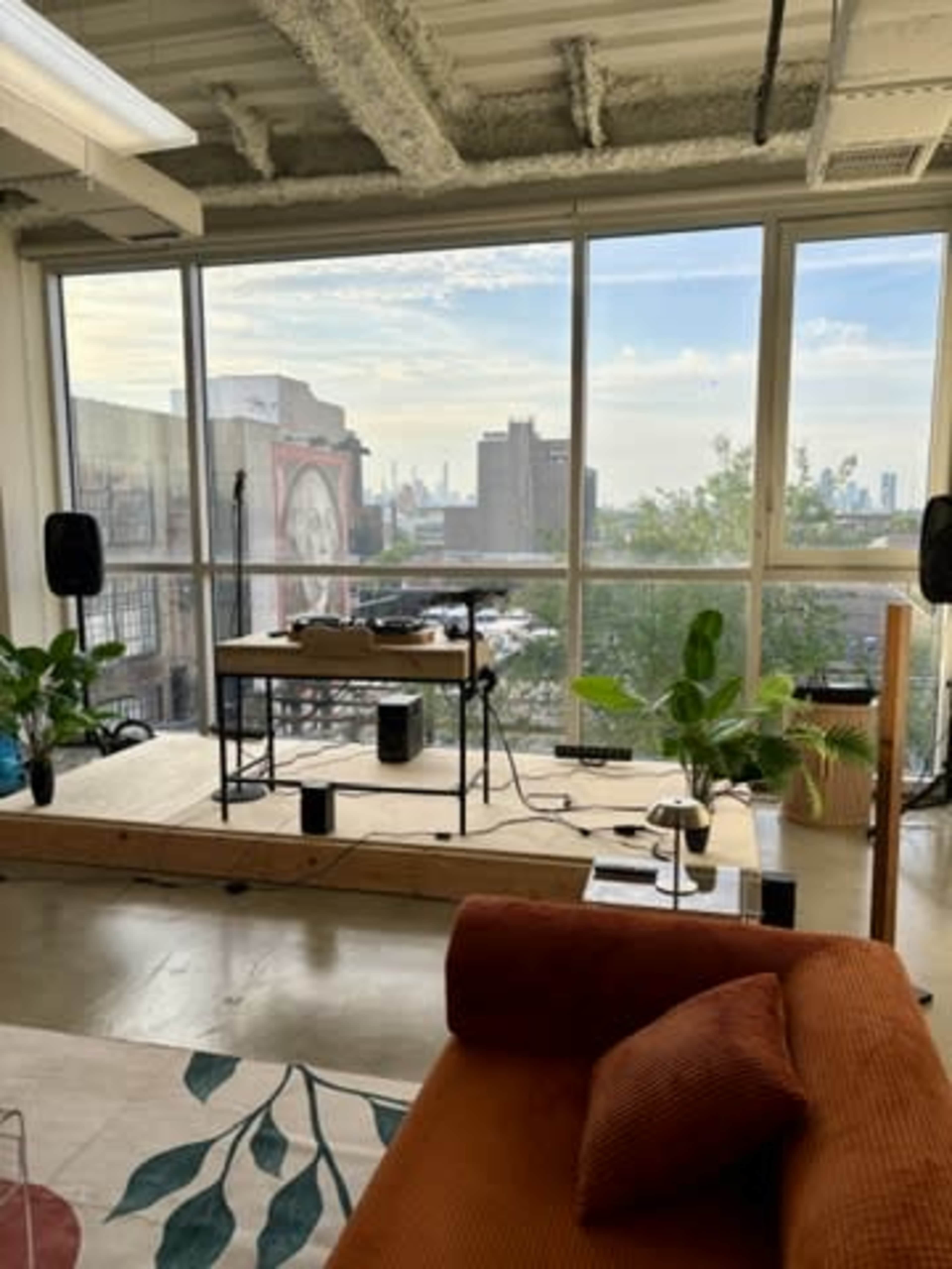 The image shows a modern interior space with a wooden platform set up for a performance, featuring plants, speakers, and a view of a city skyline through large windows.