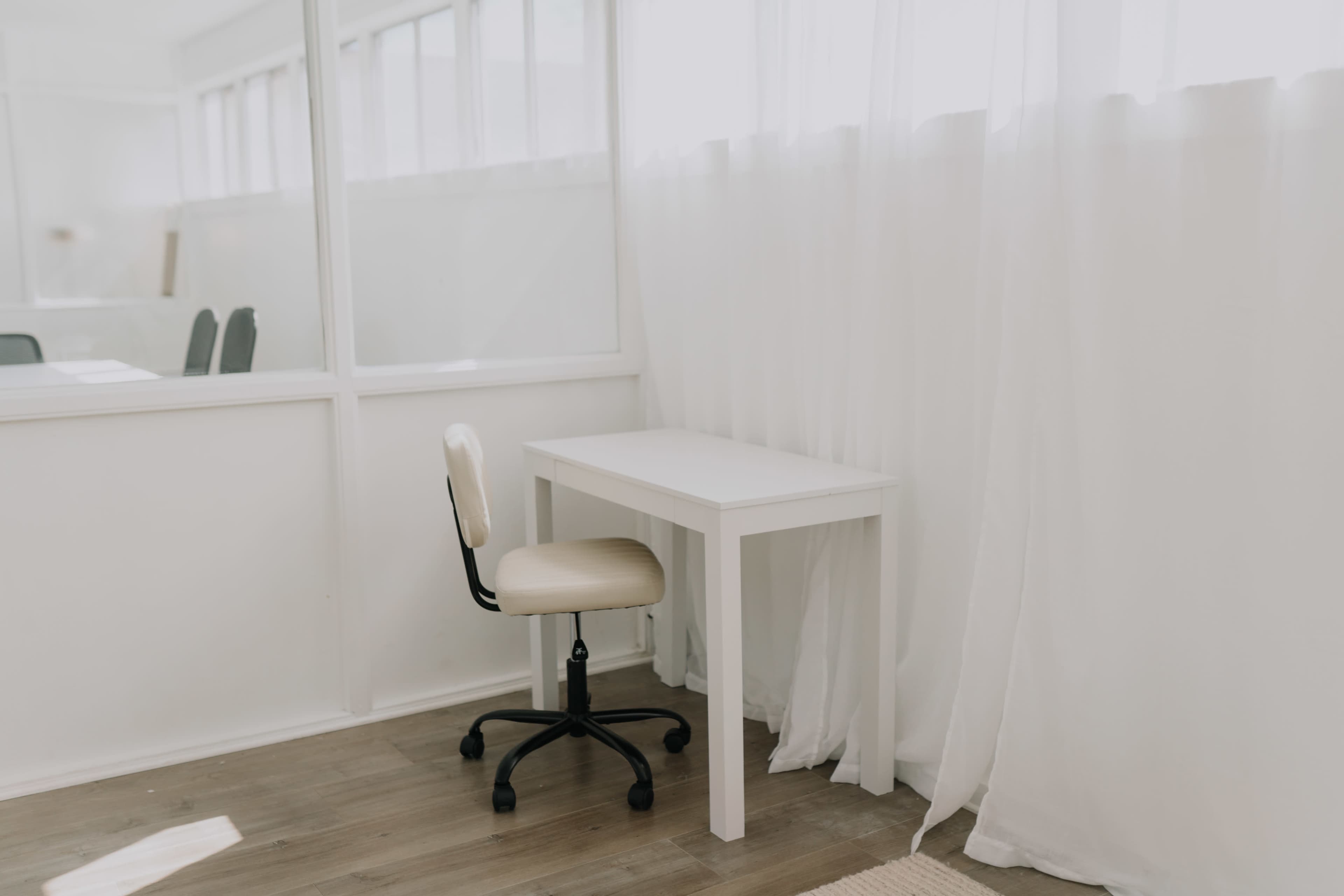 A white desk with a beige rolling chair is positioned next to sheer curtains in a bright, minimalistic room.