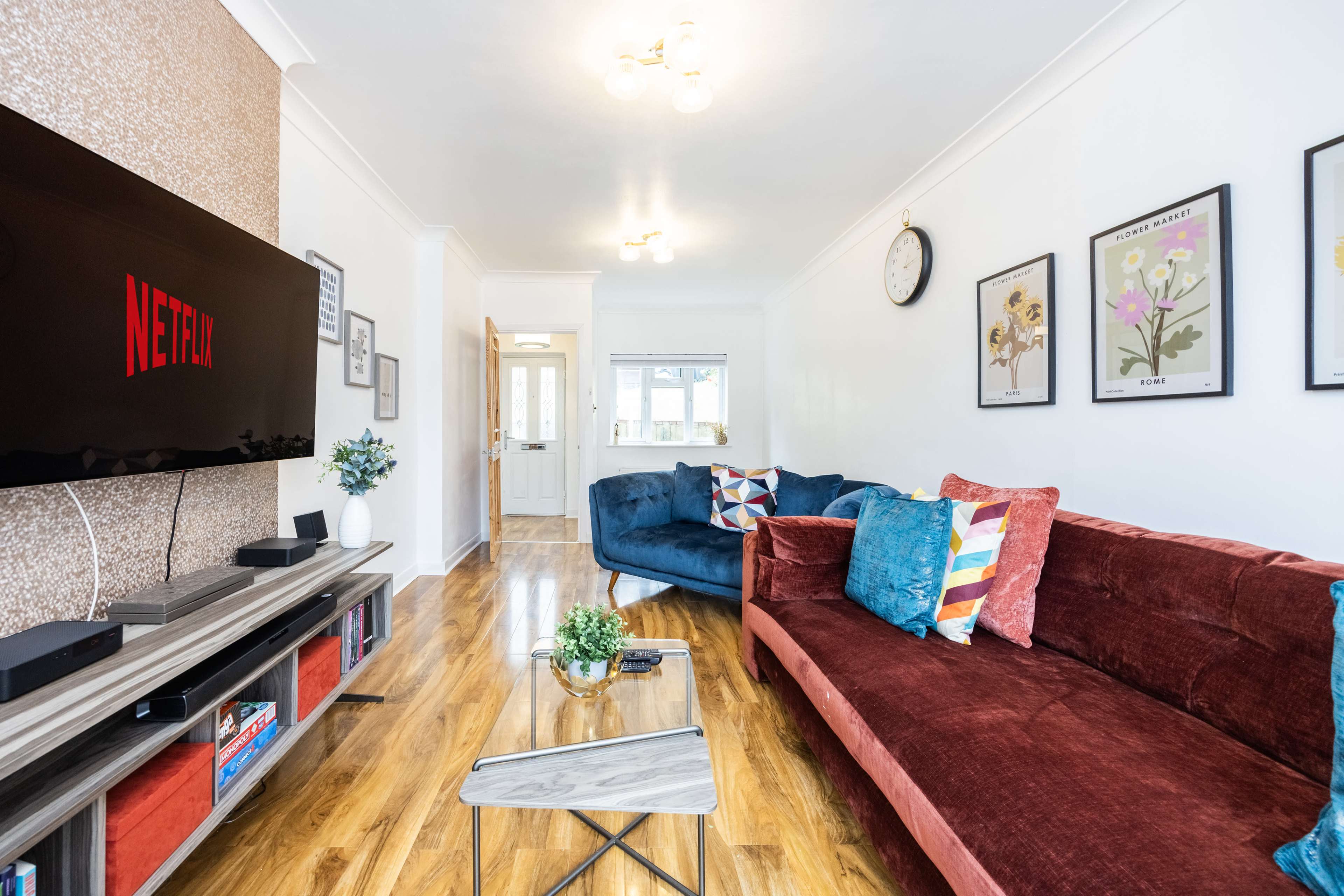 The living room features a television displaying the Netflix logo, a red sofa, a blue couch, a coffee table, and framed artwork on the walls.