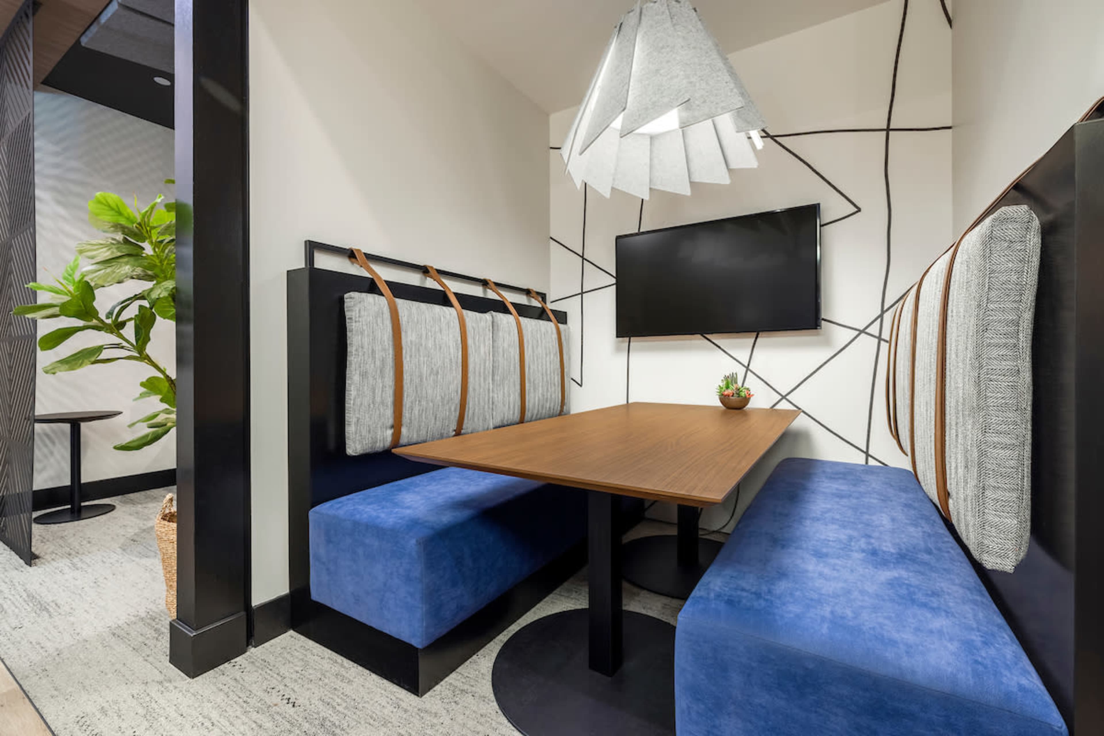 A modern meeting booth with a wooden table, blue upholstered benches, a wall-mounted television, and a decorative plant in a corner.