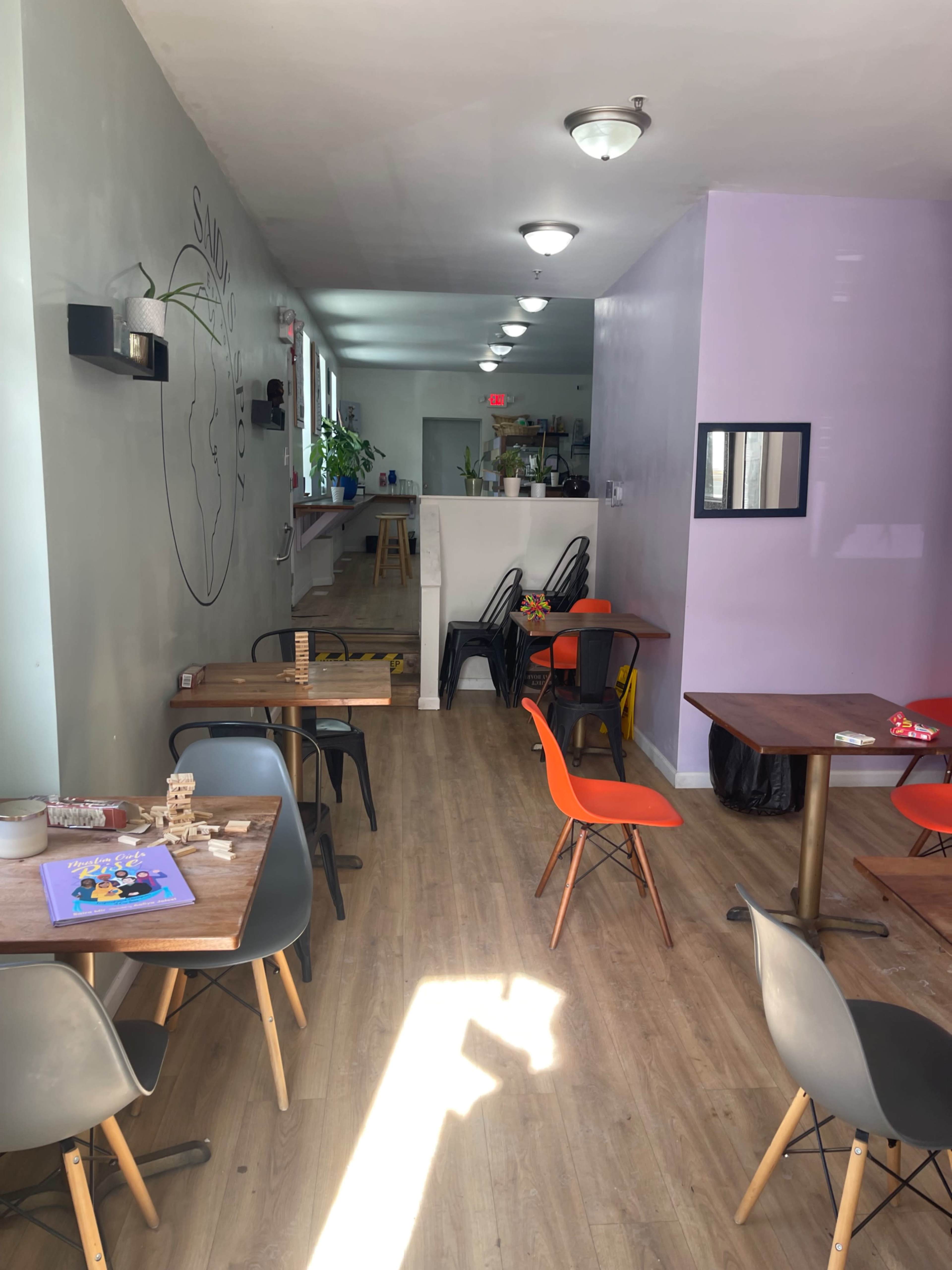The image shows a light-filled cafe with wooden floors, seating arrangements consisting of wooden and plastic chairs, and a wall featuring a simple mural.