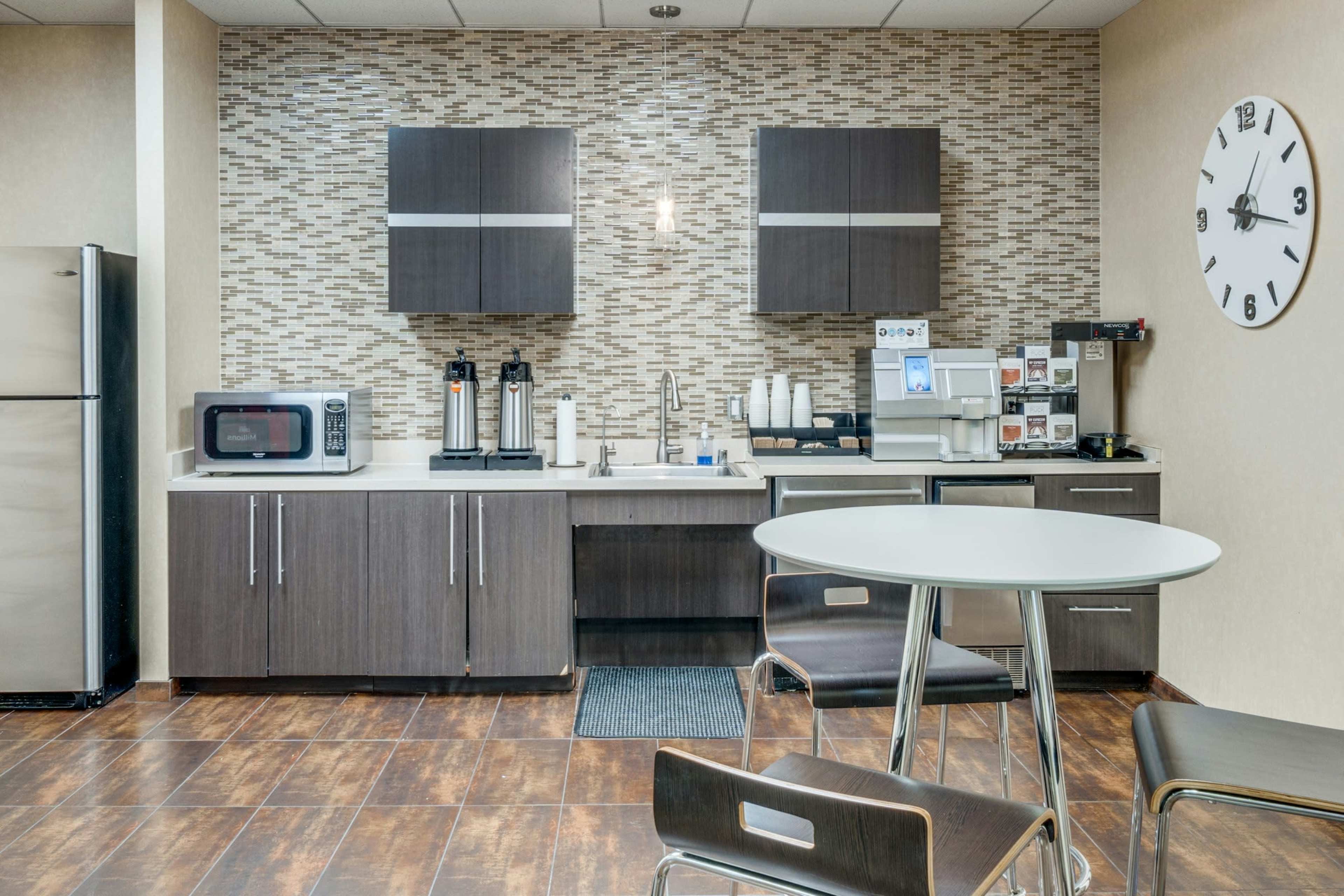 The image shows a modern kitchenette with dark cabinets, a refrigerator, a microwave, coffee dispensers, and a small round table with metal chairs.