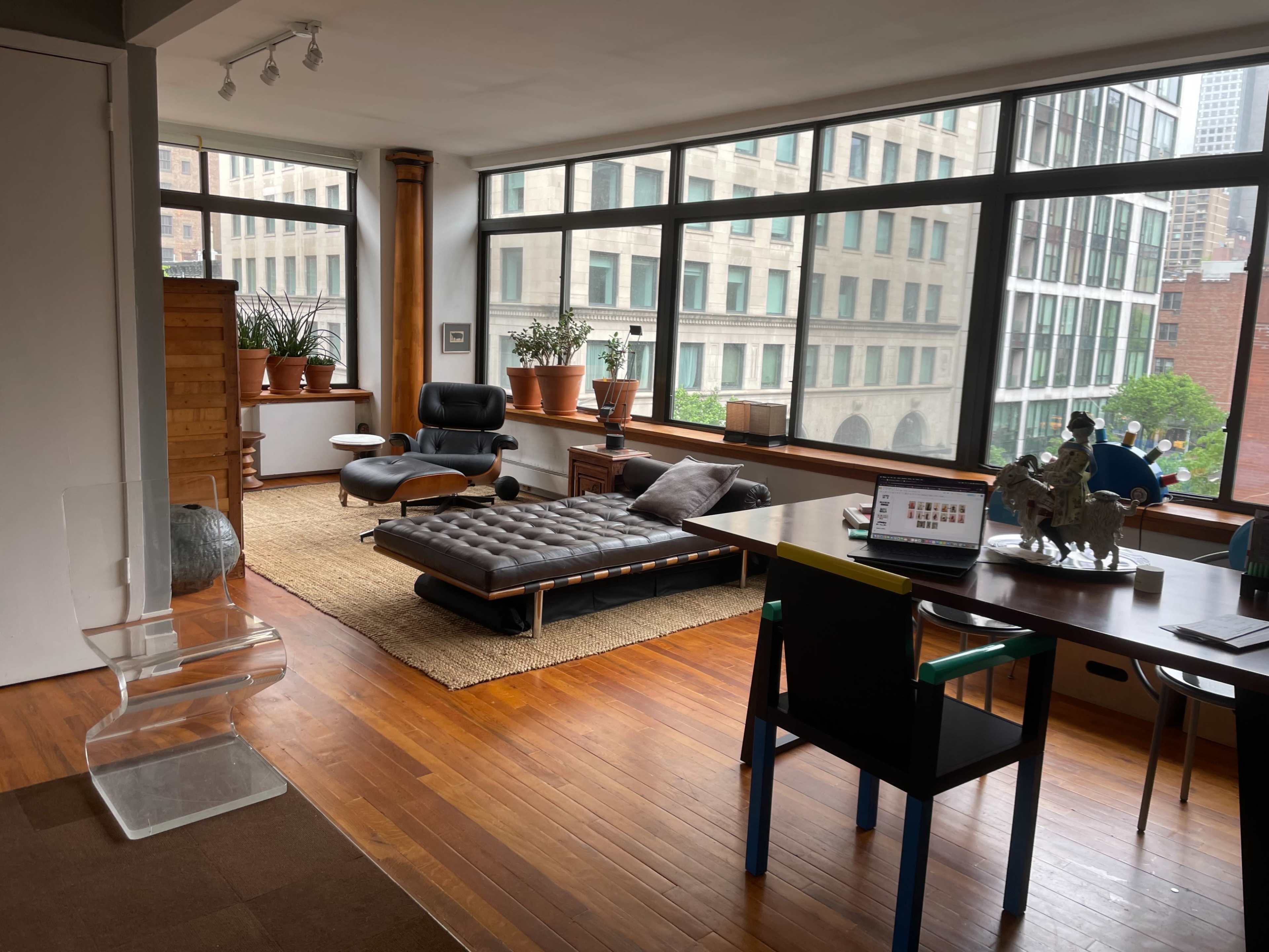 The image shows a modern living space with large windows, wooden flooring, a sectional sofa, a lounge chair, and various indoor plants.