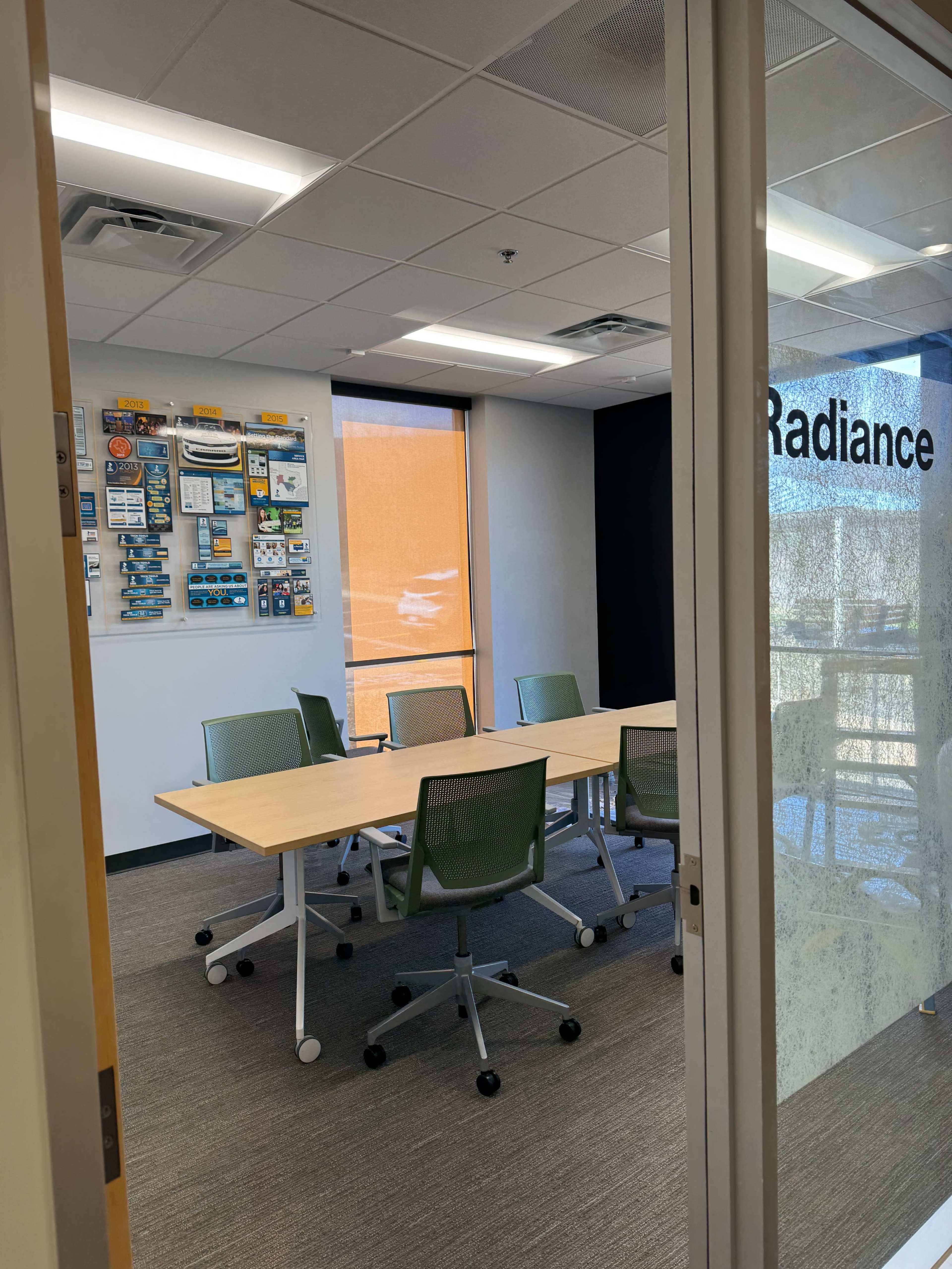 A conference room features a long wooden table surrounded by green chairs, with a display board on the wall and a large window allowing natural light in.