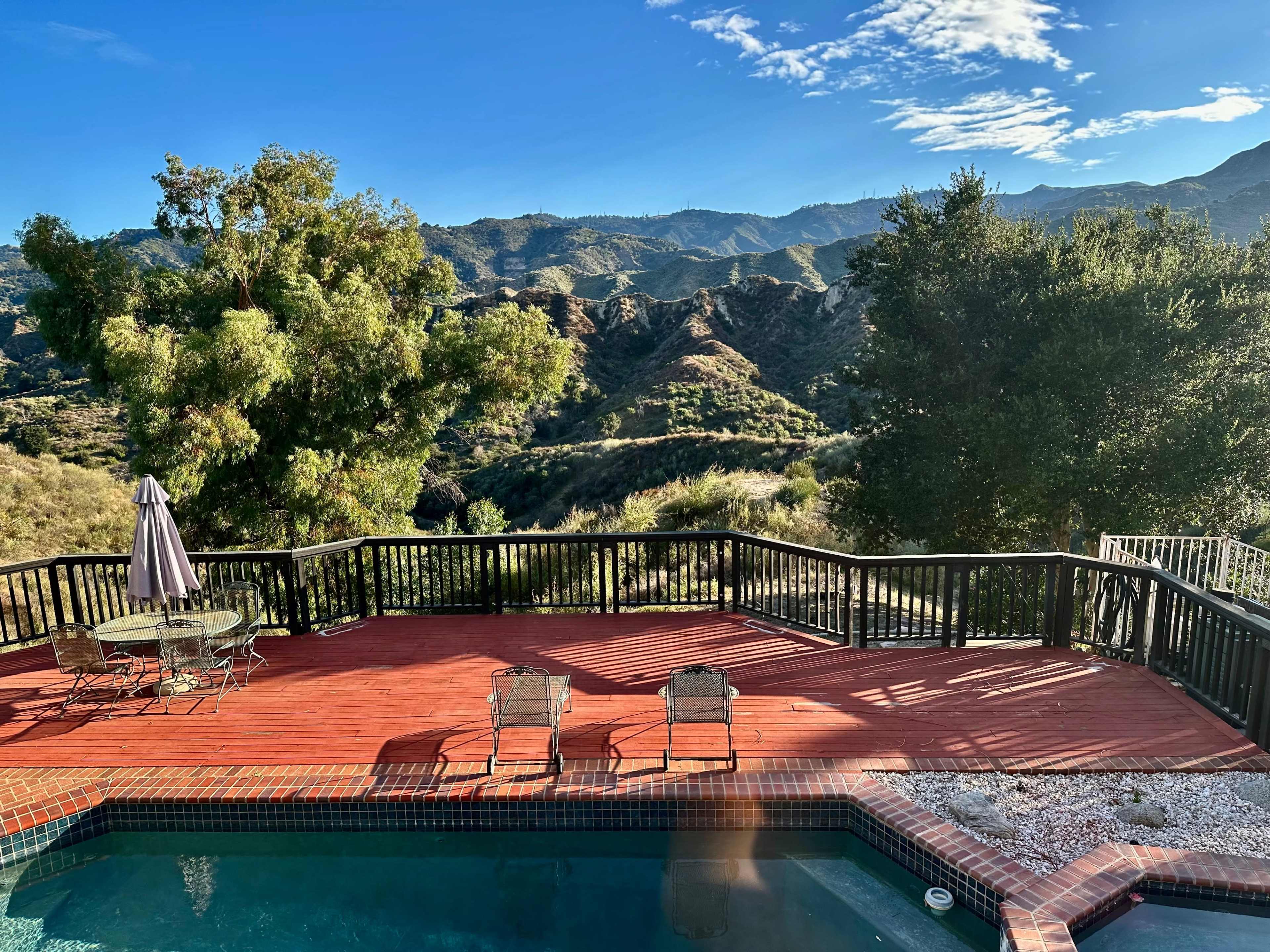 A wooden deck with two chairs and a table overlooks a pool and a mountainous landscape under a blue sky with scattered clouds.