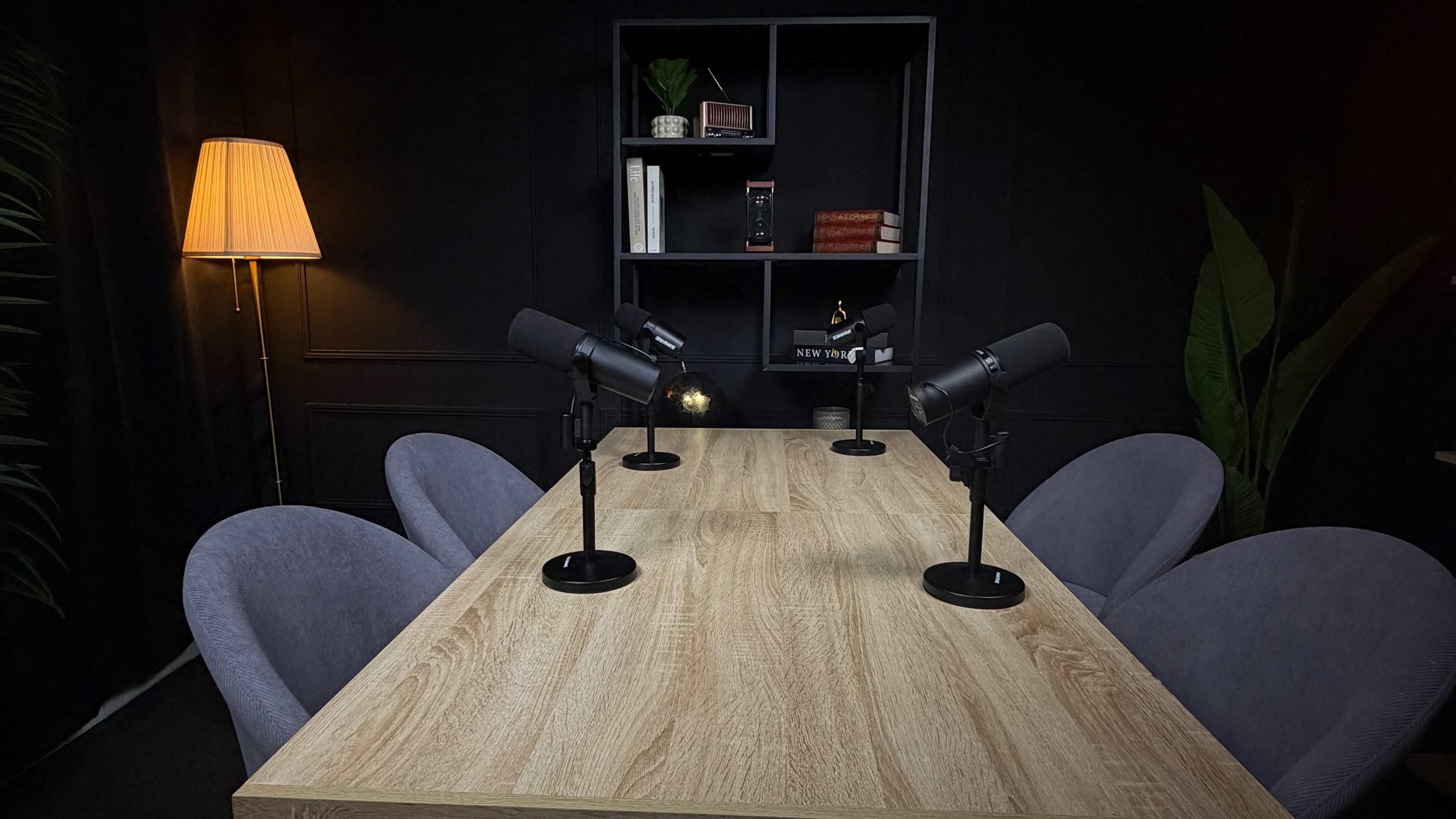 A wooden table surrounded by four microphone-equipped chairs is set in a dimly lit, modern studio with a black backdrop and shelves displaying decorative items.