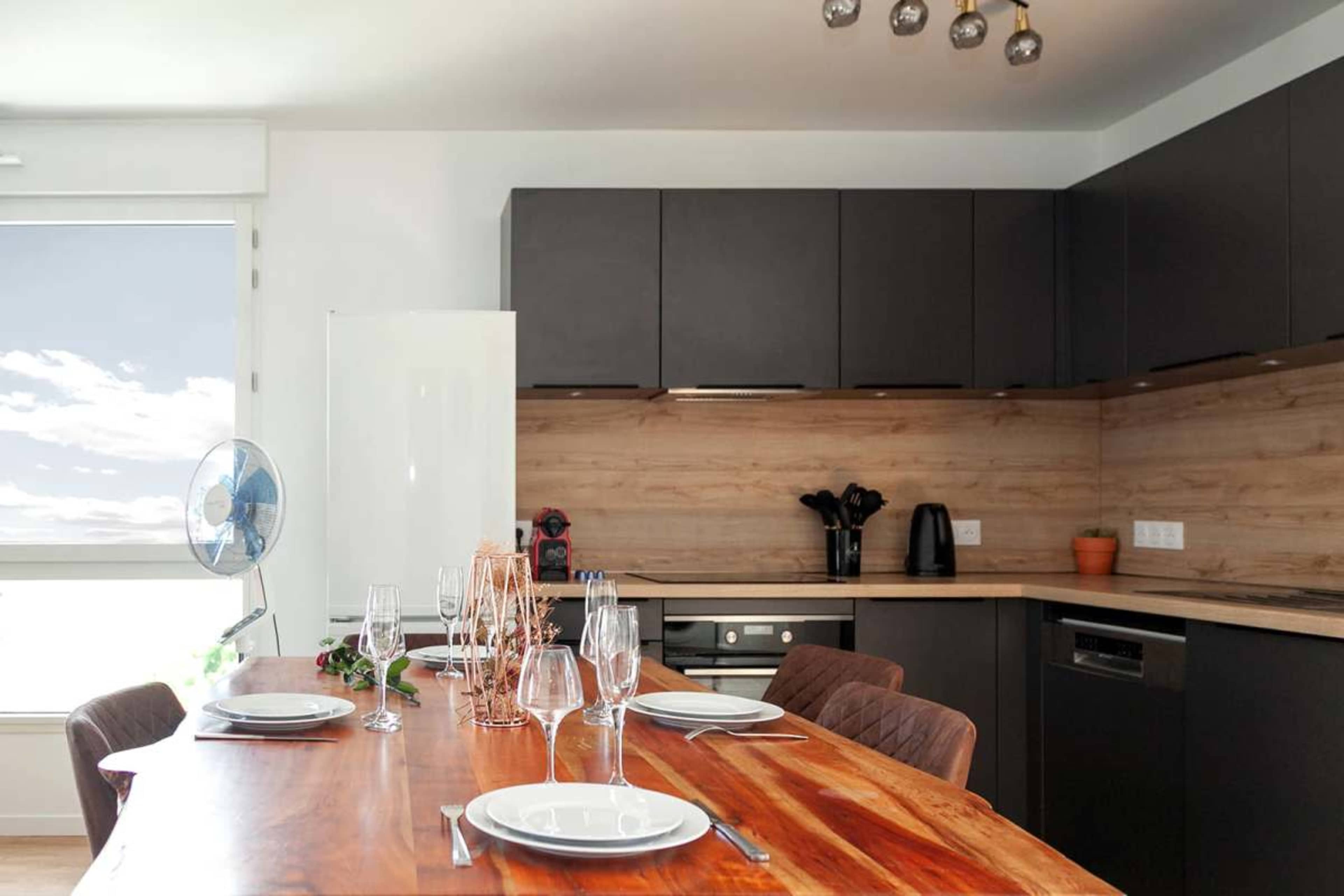A modern kitchen features dark cabinetry, a wooden countertop, and a dining table set with plates and glasses.