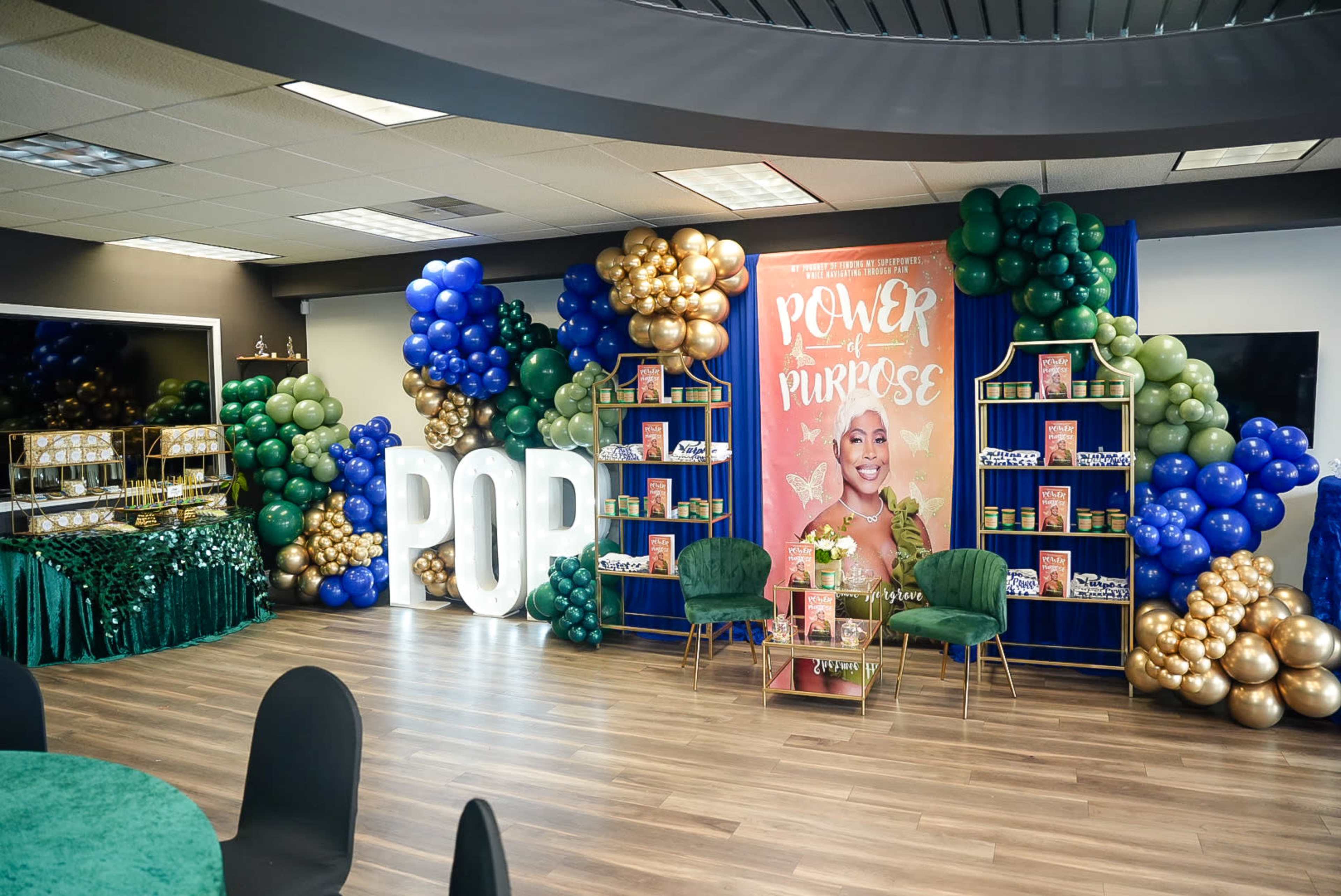 The image shows a decorated event space featuring vibrant balloon arrangements, a colorful backdrop with the words "Power Purpose," and display shelves with various products.
