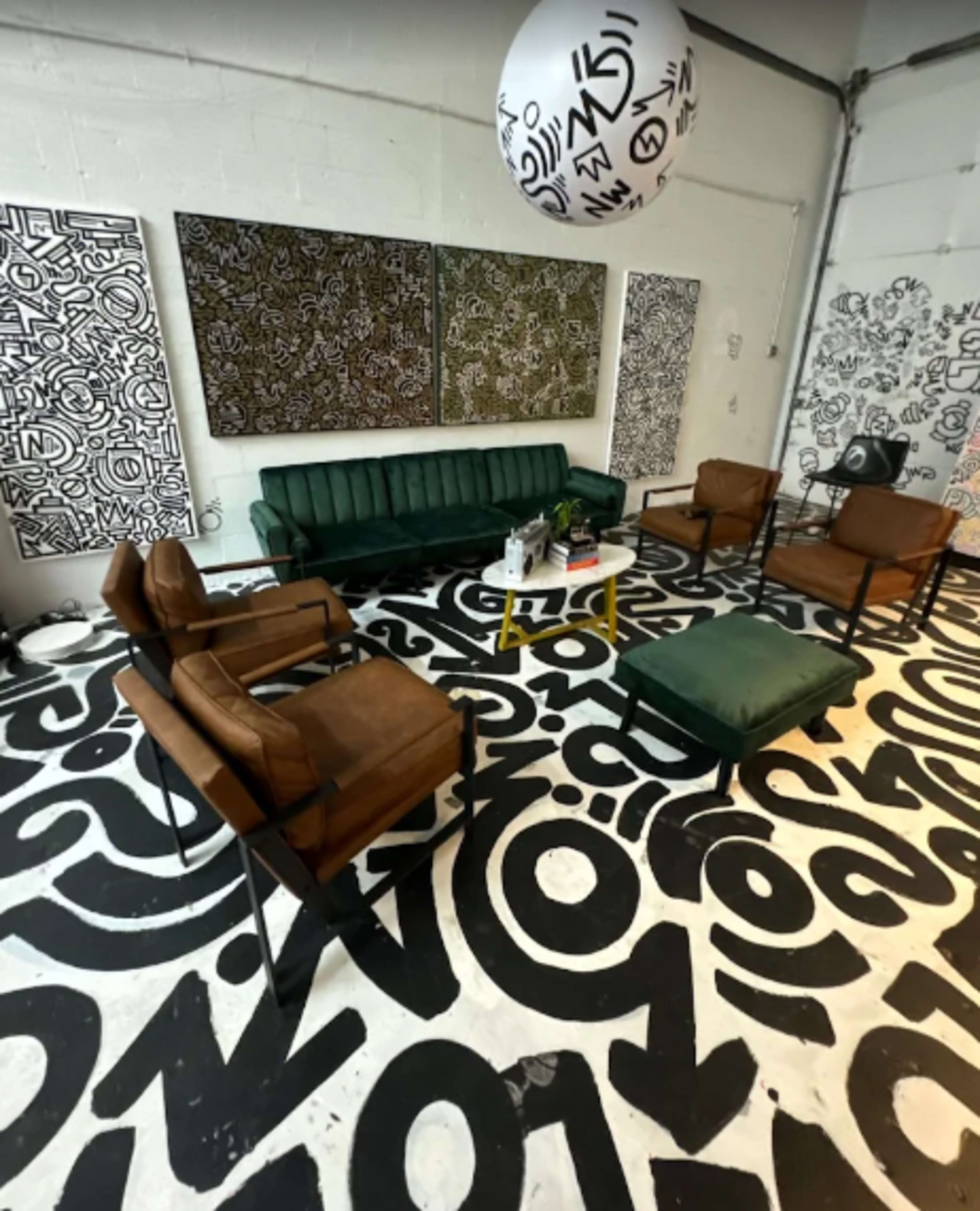 The image shows a modern lounge area with a green sofa, several brown chairs, a white table, and walls and floor decorated with bold, abstract black and white patterns.