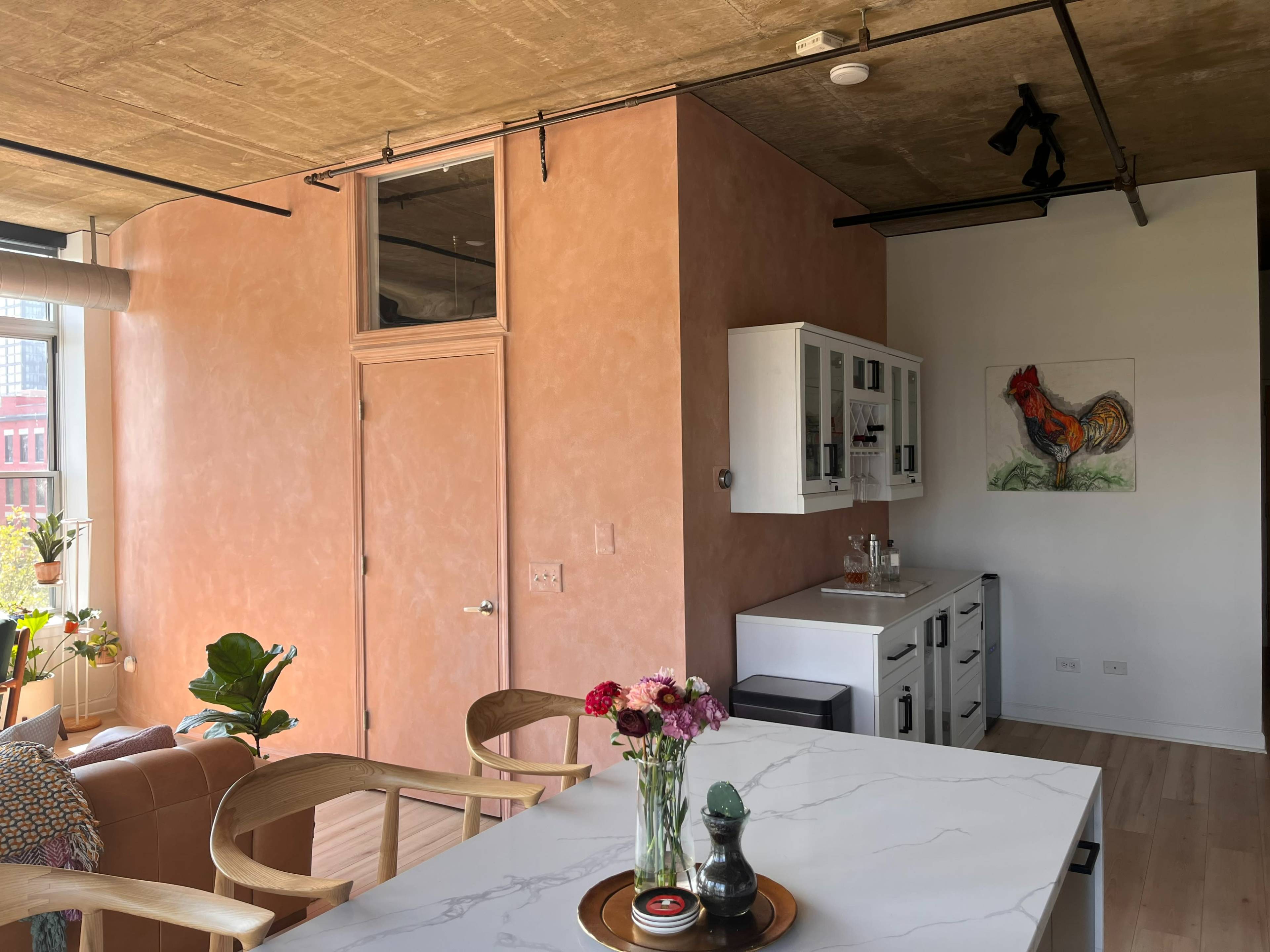 A modern kitchen and dining area with a light-colored marble table, a pink-hued wall, and a large window letting in natural light.