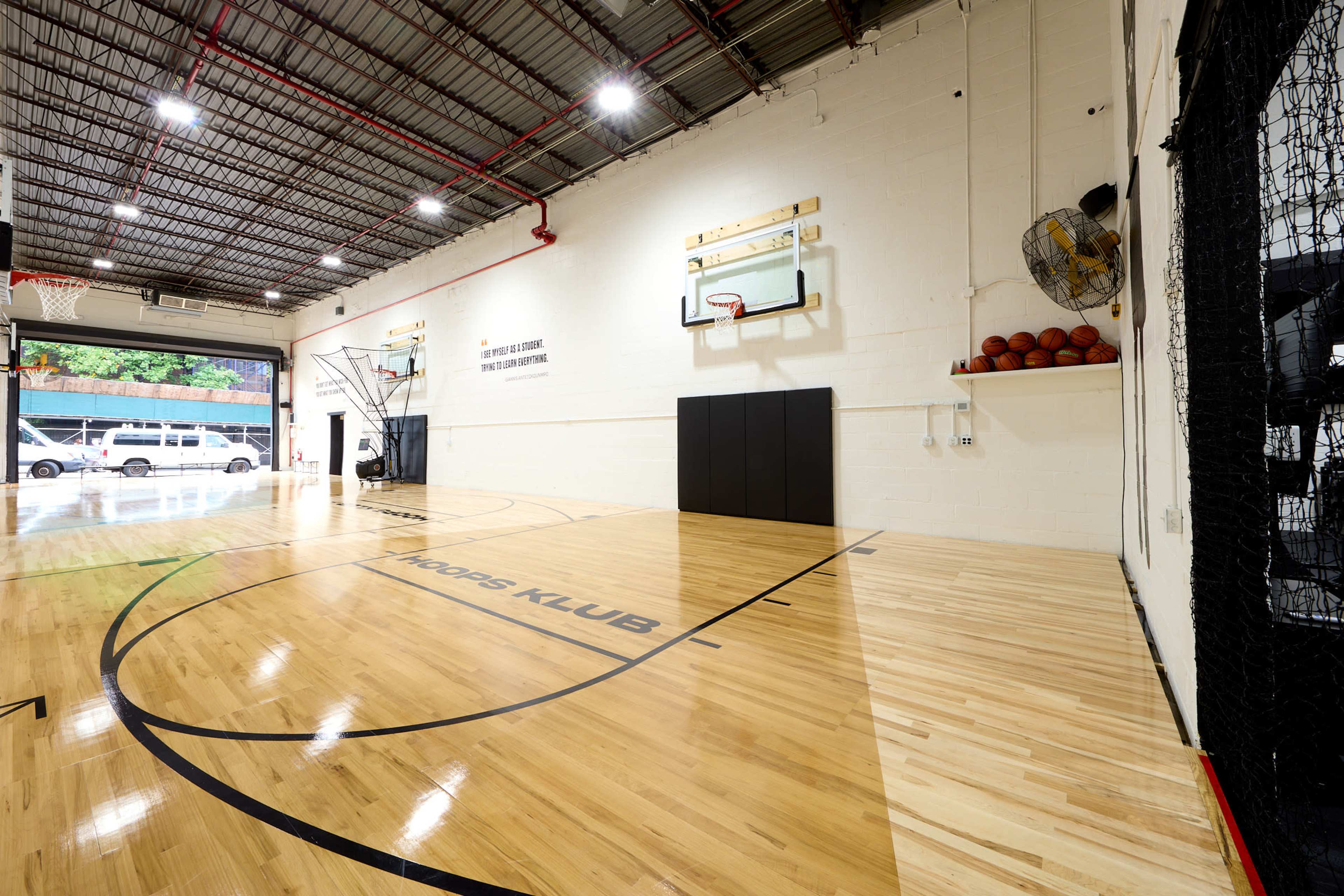 The image shows an indoor basketball court with polished wooden floors, two basketball hoops, and basketballs stacked in a corner.