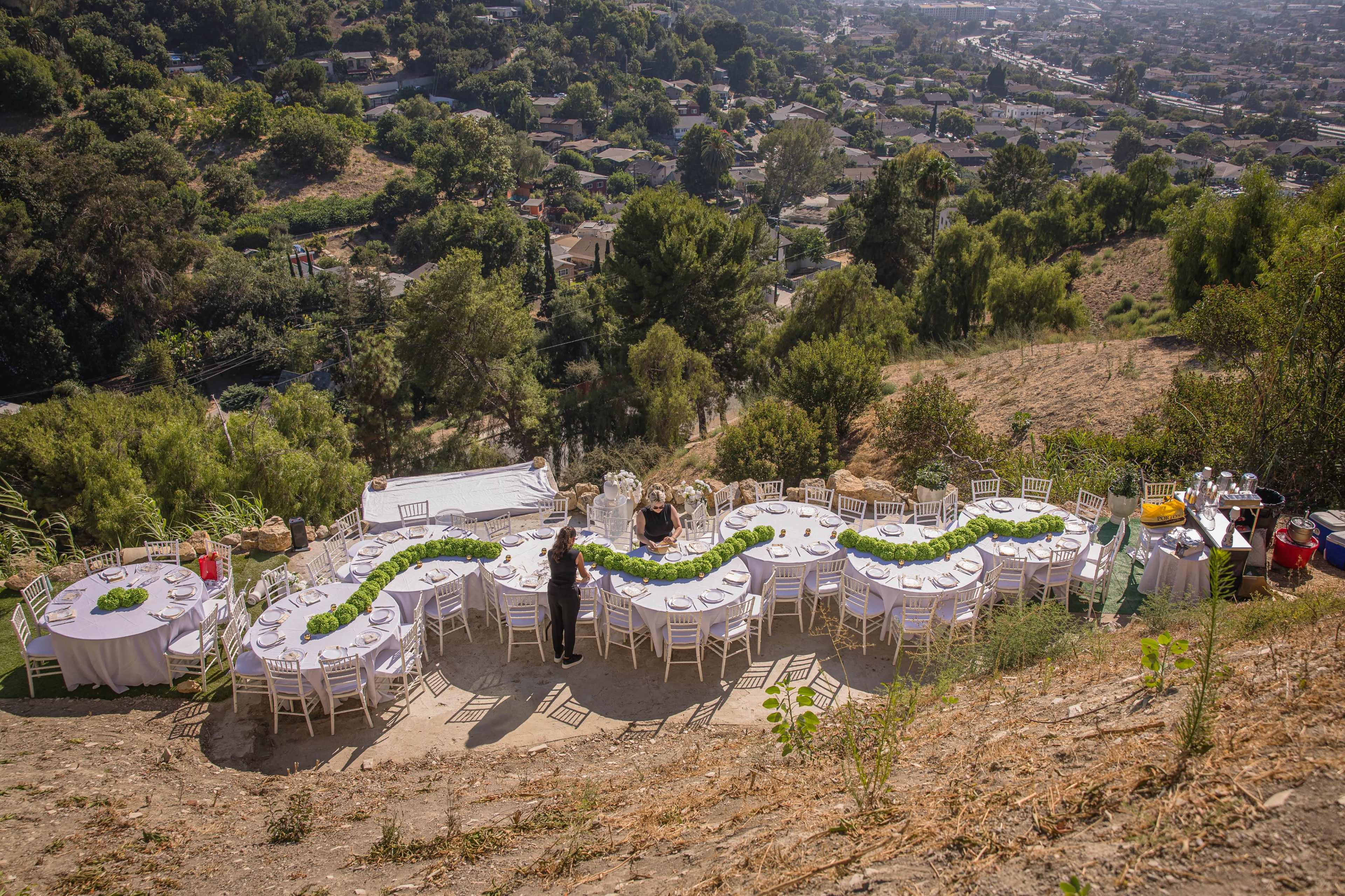 A long outdoor dining table is set up on a hillside, surrounded by greenery and overlooking a valley with houses below.