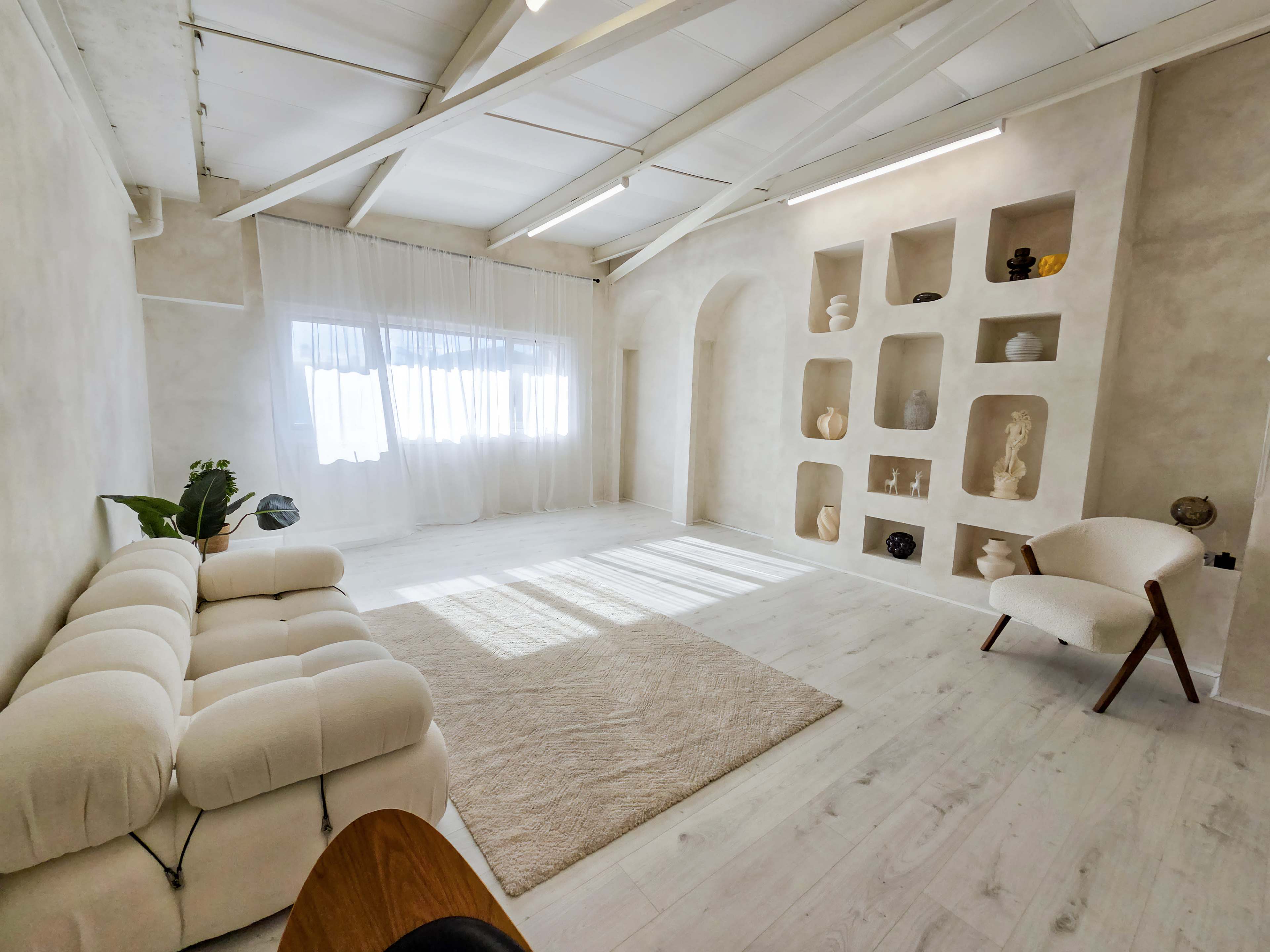 The room features a light-colored, minimalist design with a plush sofa, a small area rug, and a shelf displaying decorative objects.
