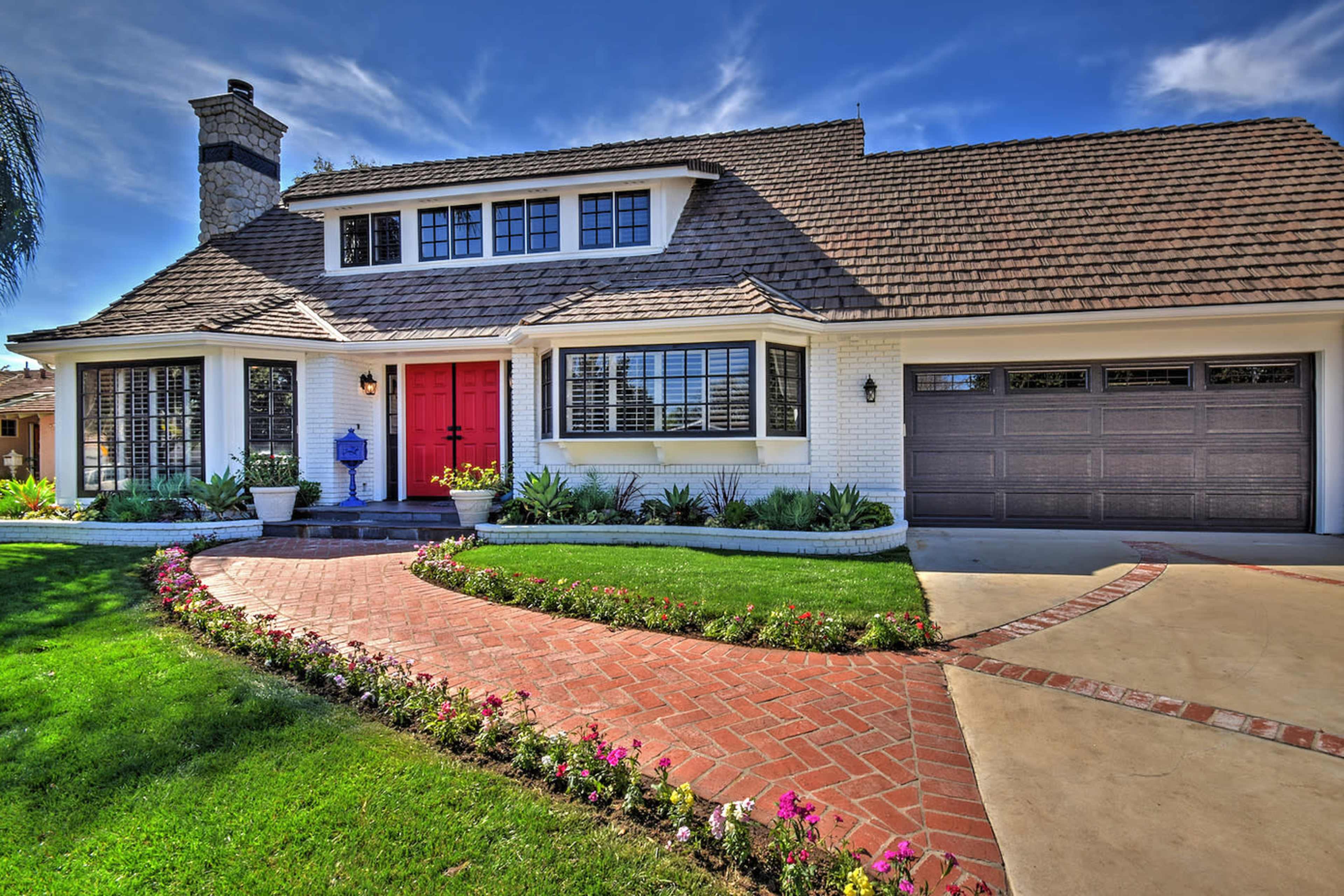 A two-story house with a red front door, large windows, and a brick walkway surrounded by a lawn and flowerbeds.