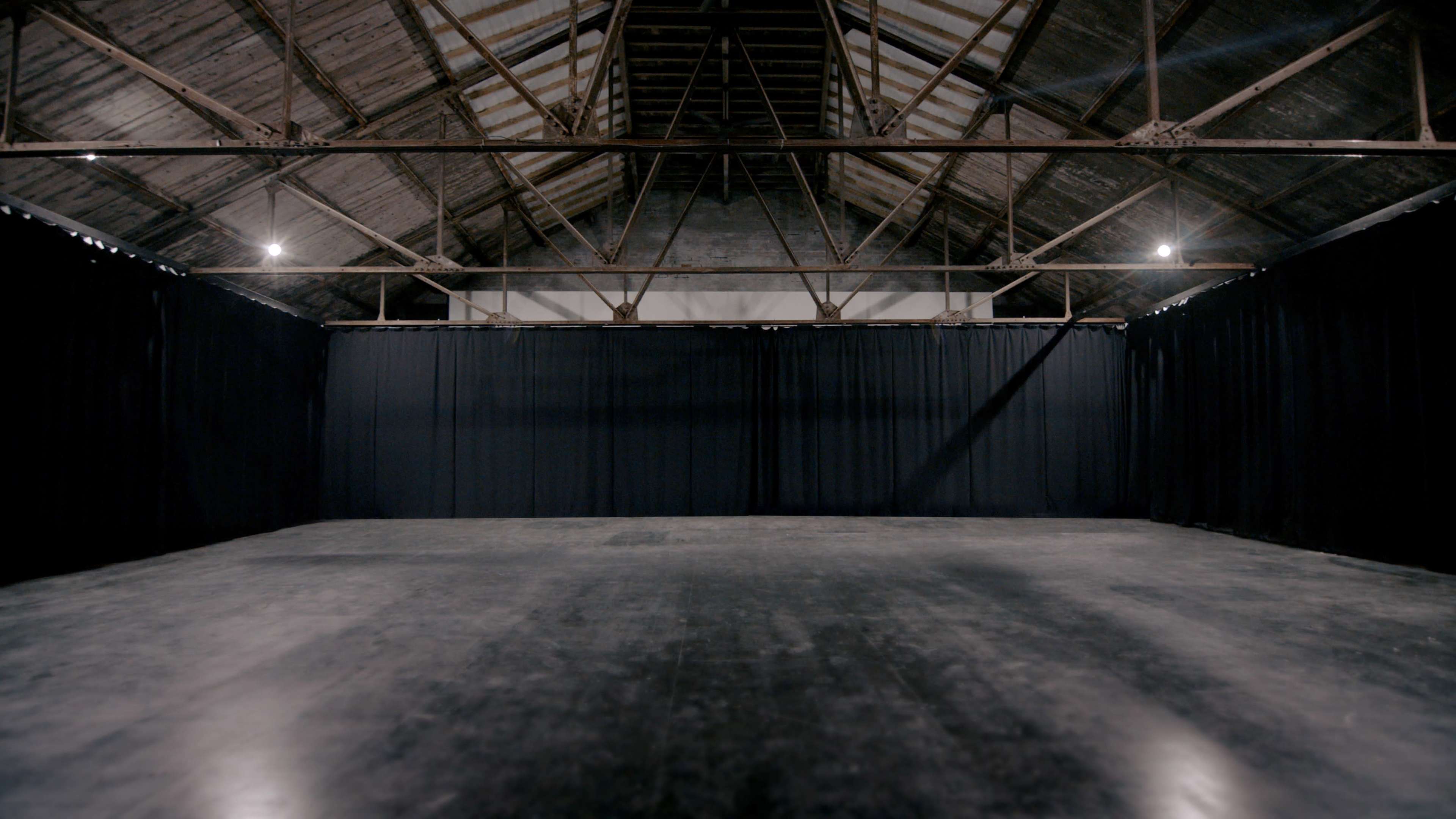 The image shows a large, empty warehouse space with a high, exposed beam ceiling and black curtains draped around the sides.