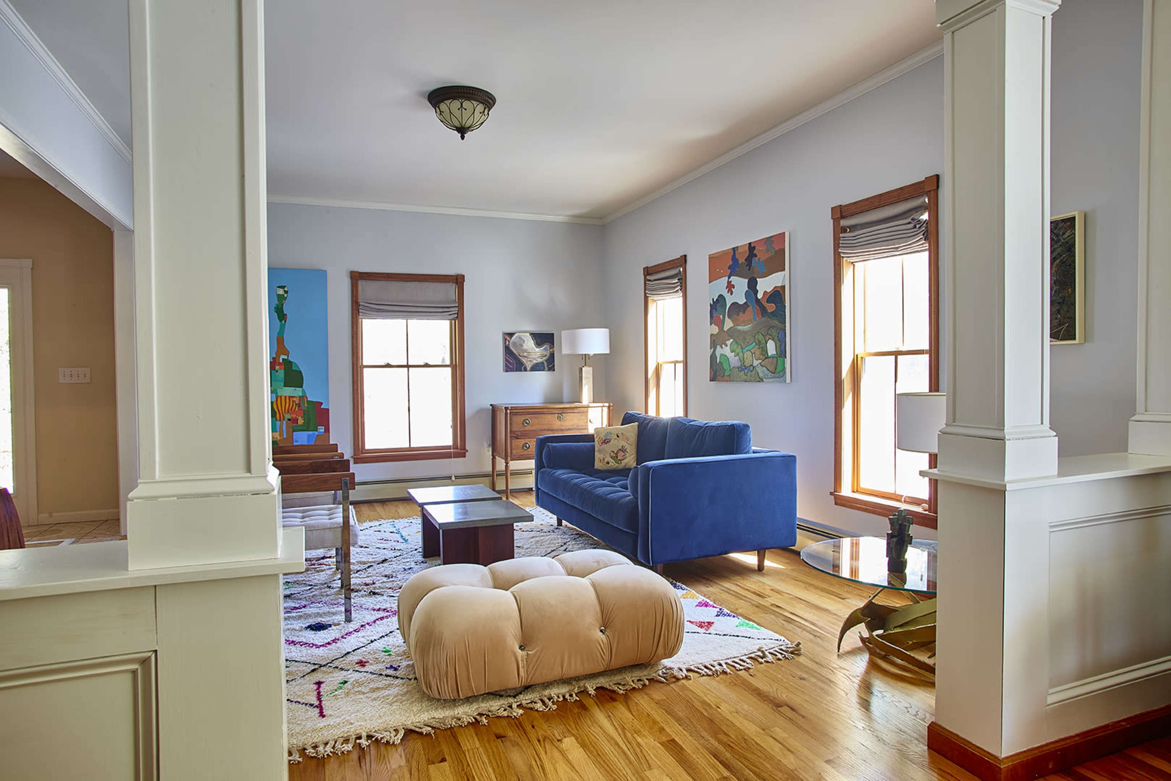 A brightly lit living room featuring a blue sofa, a wooden chest, and two large windows with blinds.