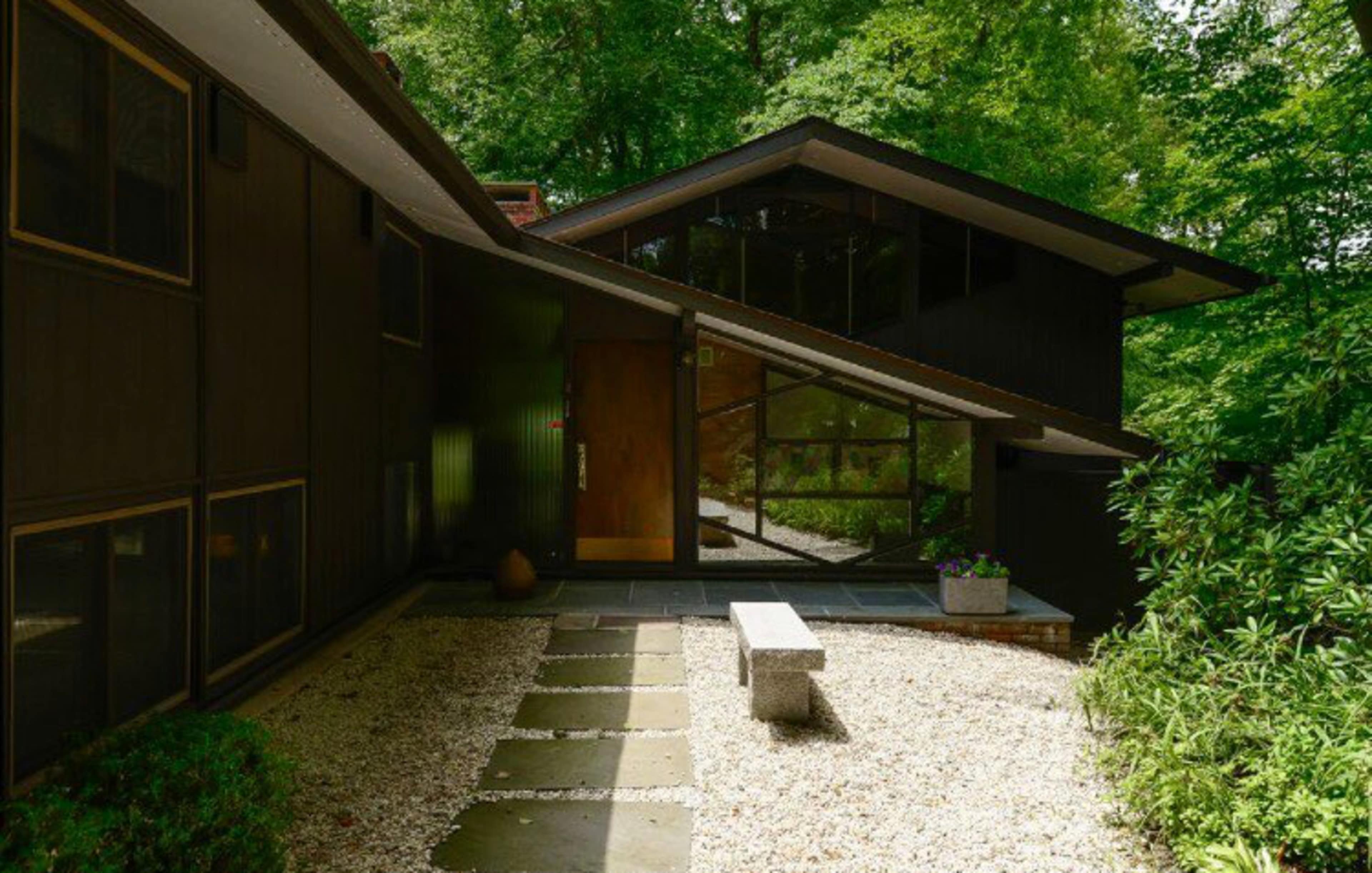 A modern house with a sloped roof and large windows, surrounded by greenery and a stone pathway leading to the entrance.