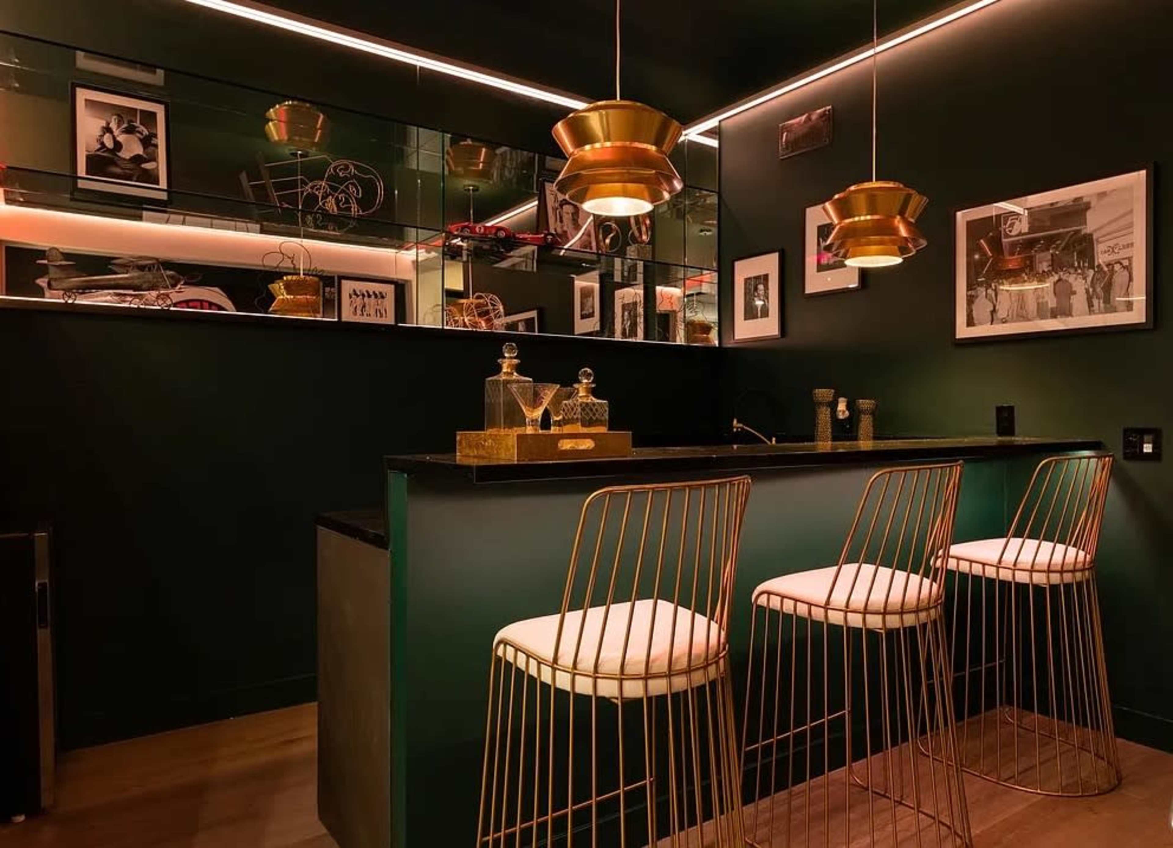 The image shows a modern bar area with a dark green wall, a sleek countertop, and three wire-frame stools, illuminated by warm pendant lights.
