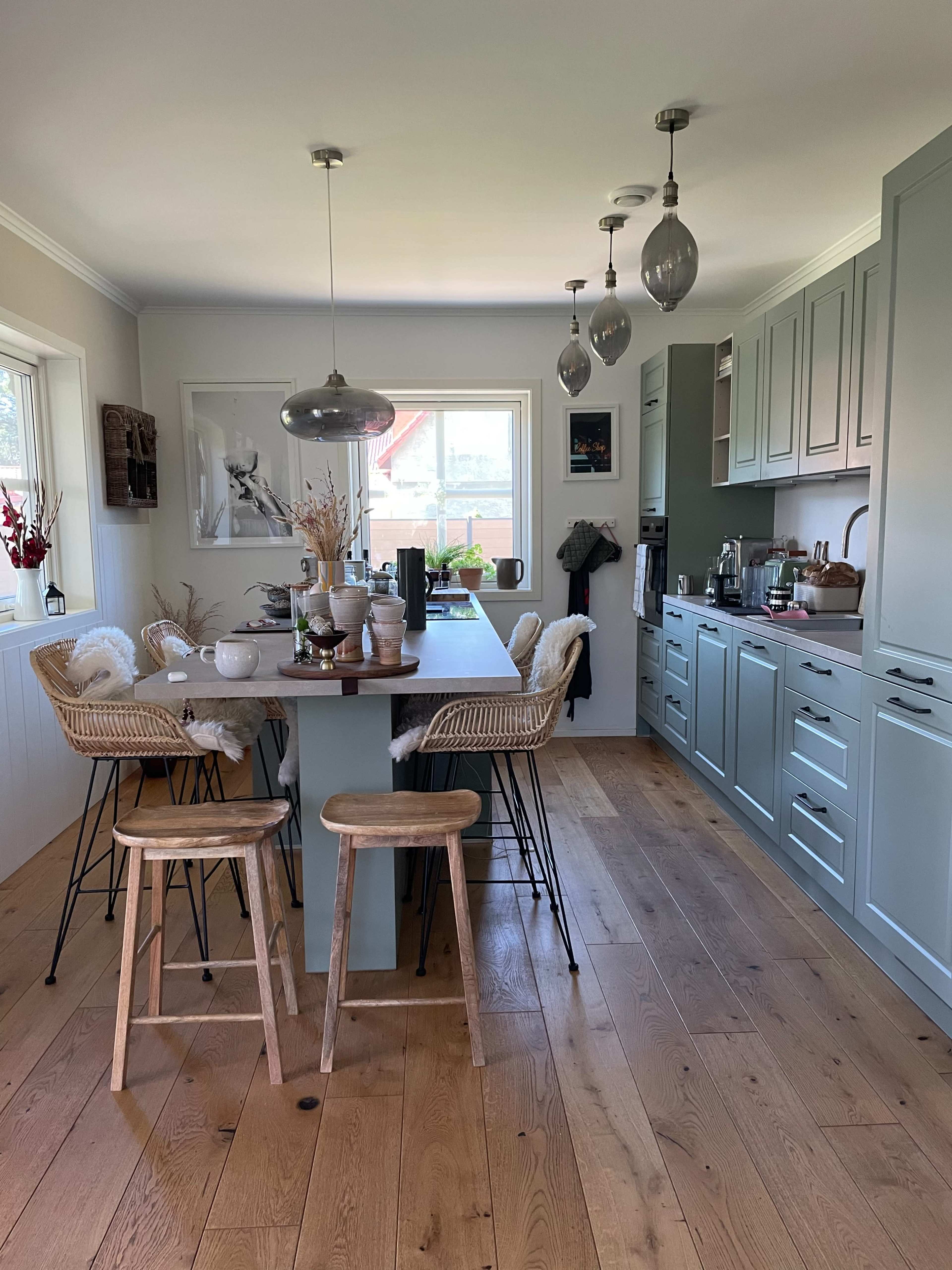 A modern kitchen features a table with three wooden stools, cabinetry with a soft blue finish, and pendant lighting above.