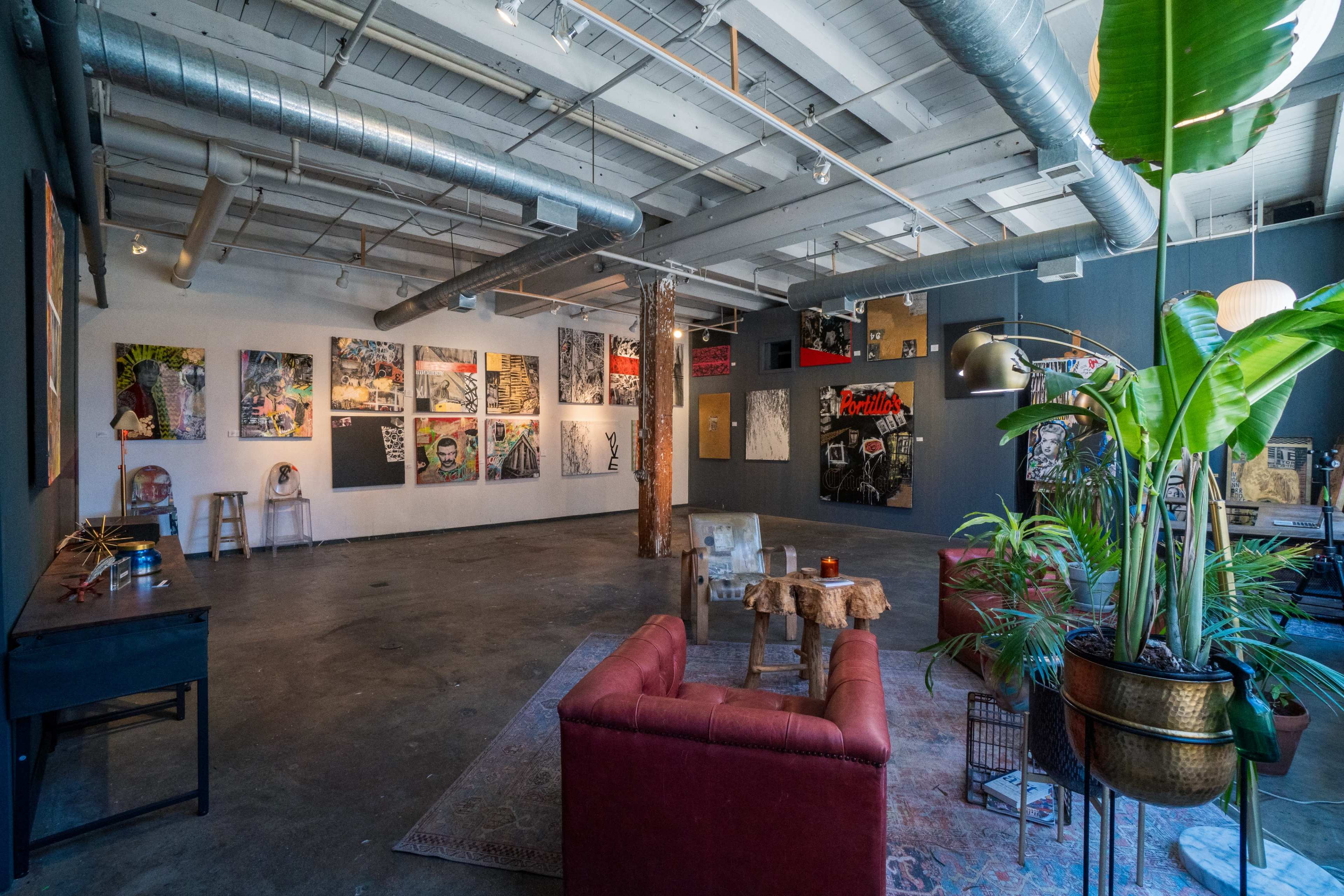 An art gallery with various paintings displayed on the walls, a seating area with red sofas, and a large potted plant in the corner.