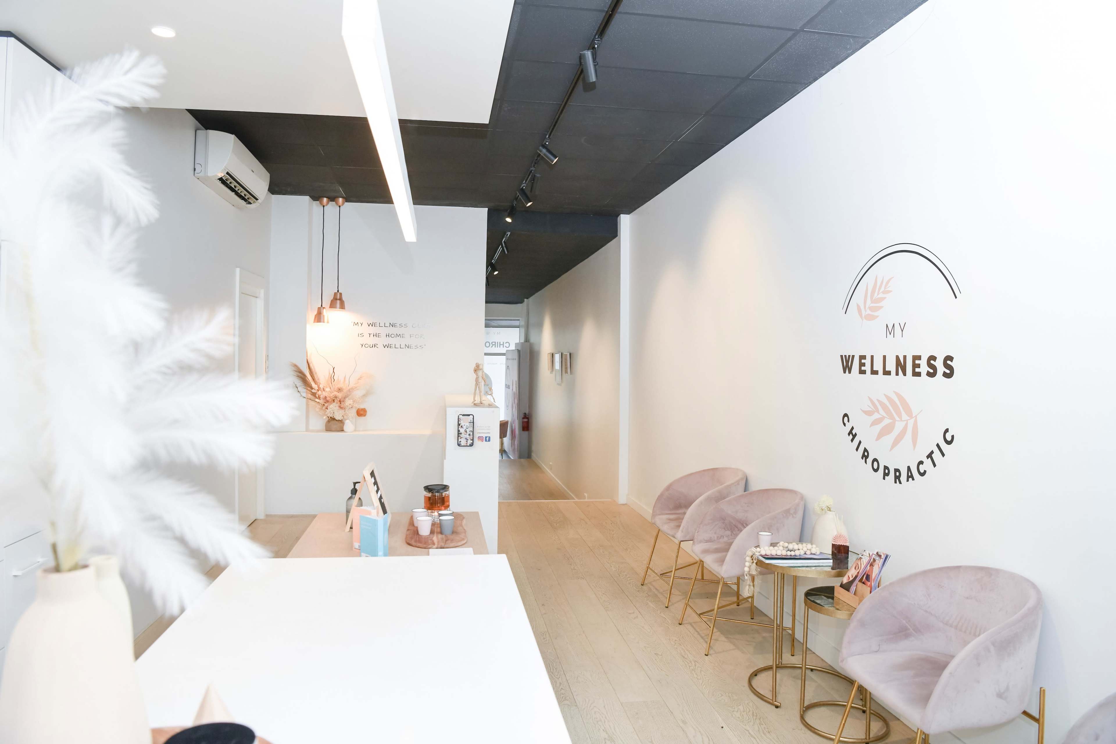 The image shows a modern chiropractic office reception area with soft pink chairs, a minimalist white counter, and decorative elements along the wall.