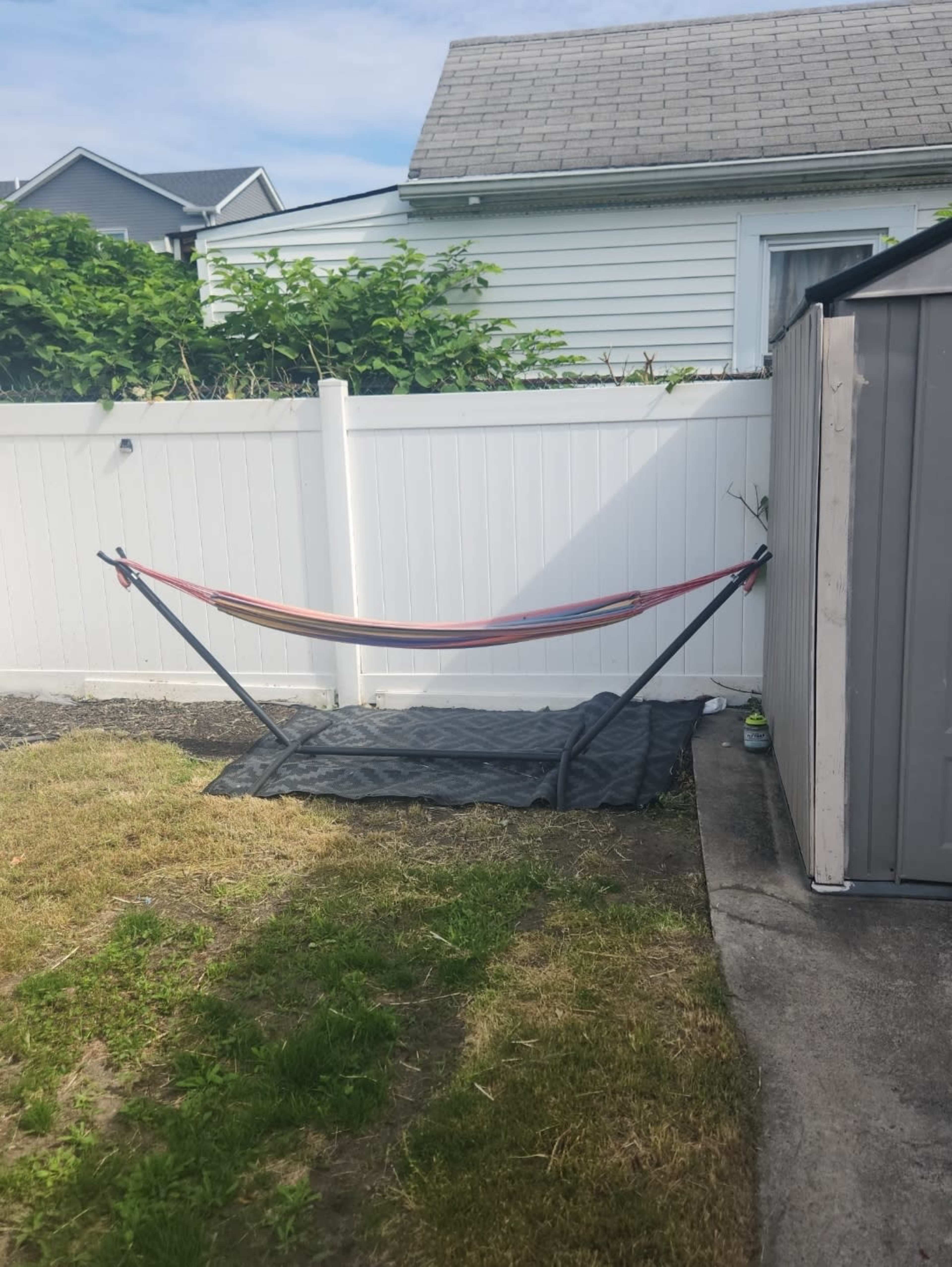 A hammock is set up on a mat beside a gray shed against a white fence in a backyard.