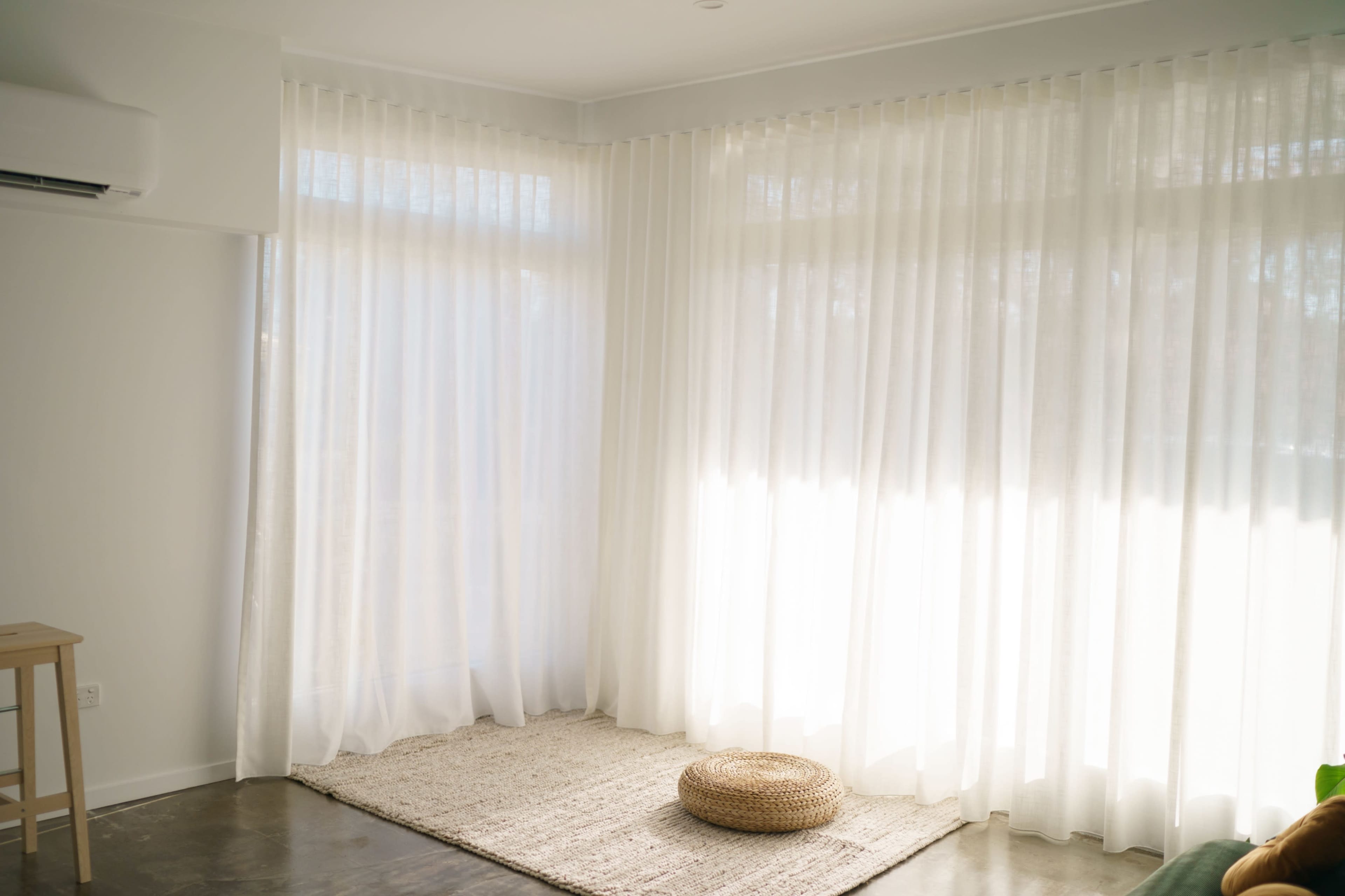 A bright room features sheer white curtains, a woven rug, and a circular pouf resting on a polished concrete floor.