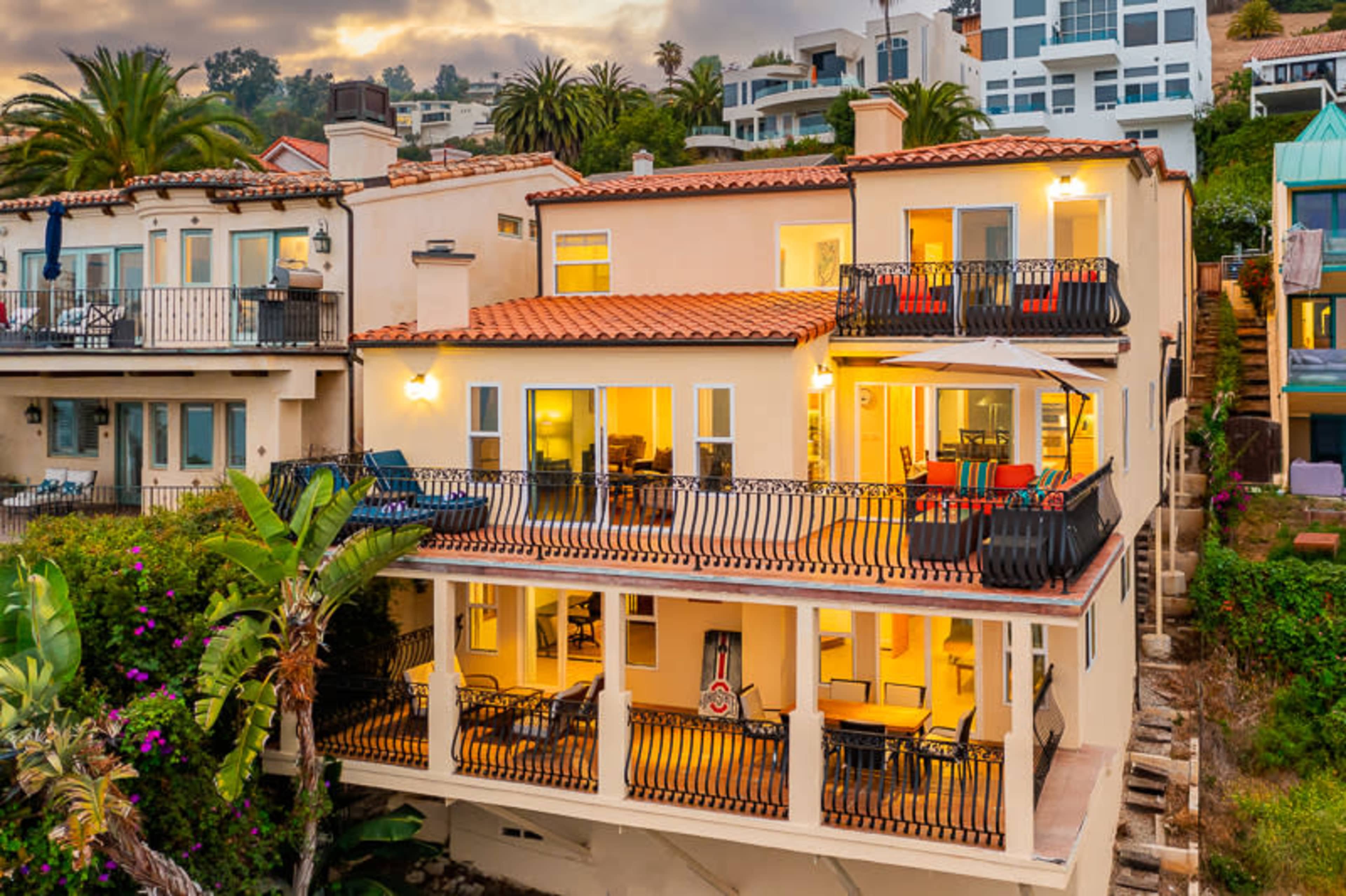 A multi-level beachfront house with a terracotta-tile roof, large balconies, and palm trees surrounding the property.
